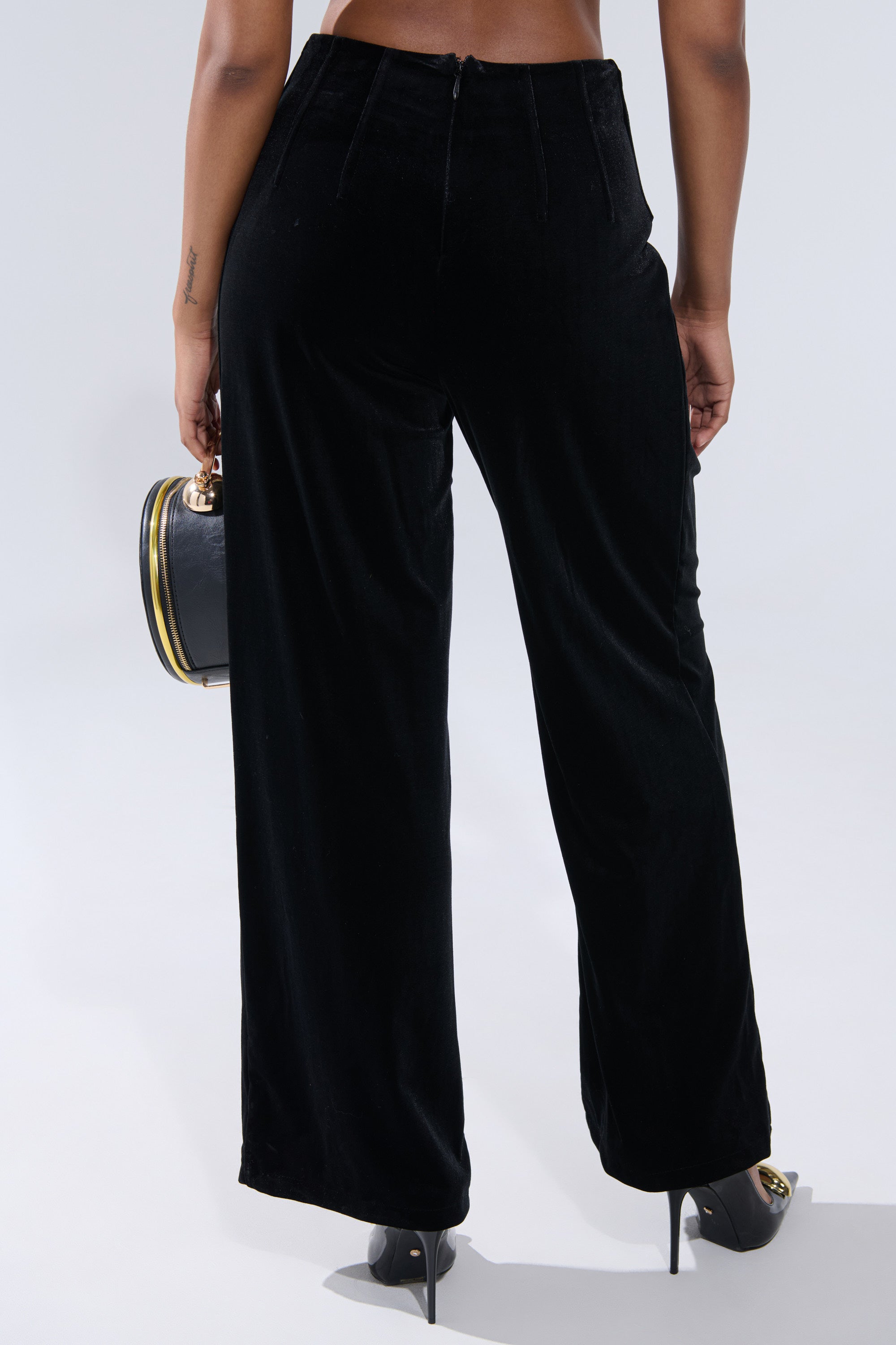 A person is seen from the back wearing SHOW ME IT VELVET TROUSER in black, styled with black heels and a black handbag with gold trim, set against a light gray background. 3