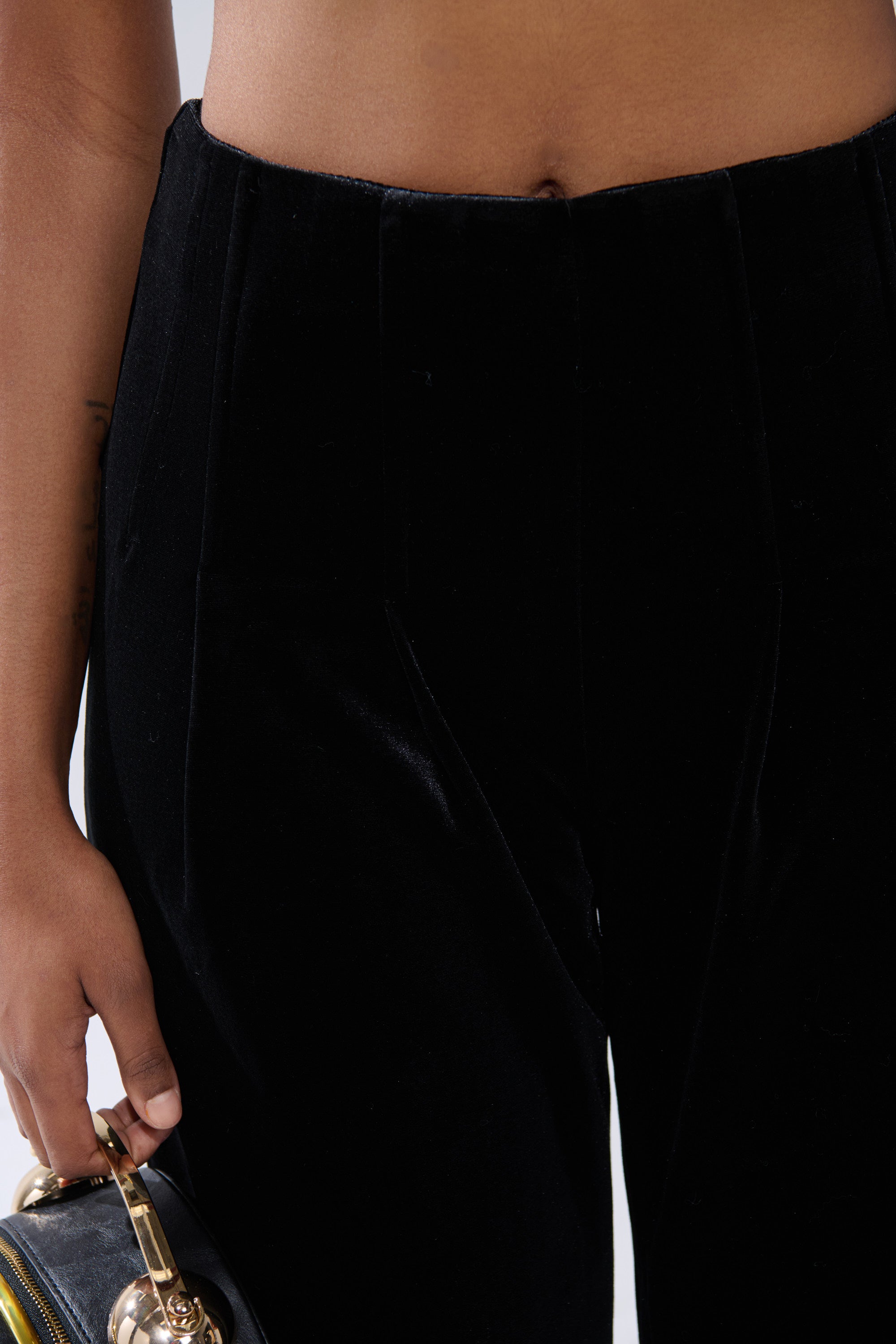 A close-up of a person wearing the SHOW ME IT VELVET TROUSER, black high-rise wide-leg velvet pants, holding a black and gold handbag in their left hand with their midriff and hand visible. 5