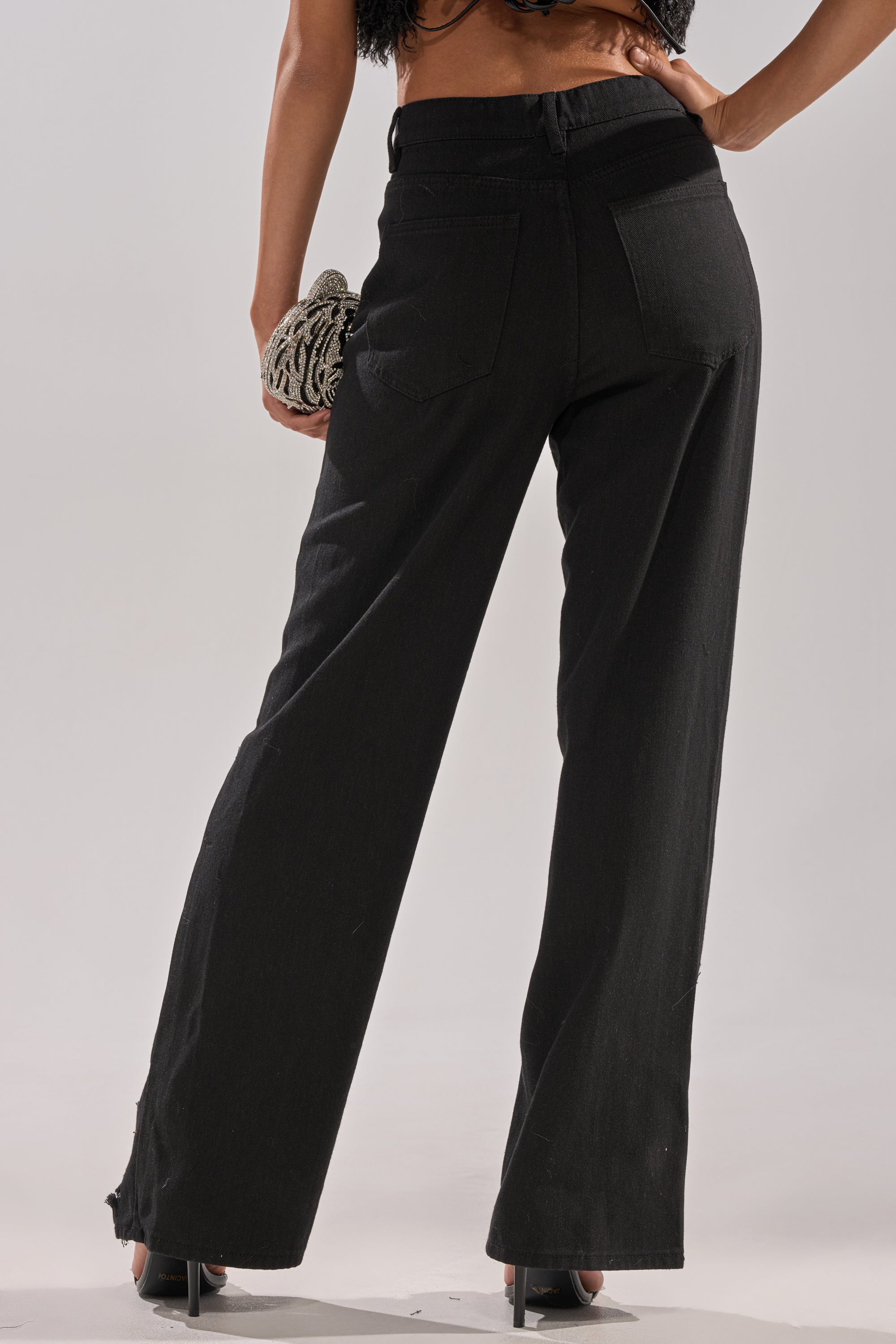 A person faces away, wearing CLARA DISTRESSED DENIM high-rise jeans with black heels, holding a textured clutch in their right hand against a plain light background.