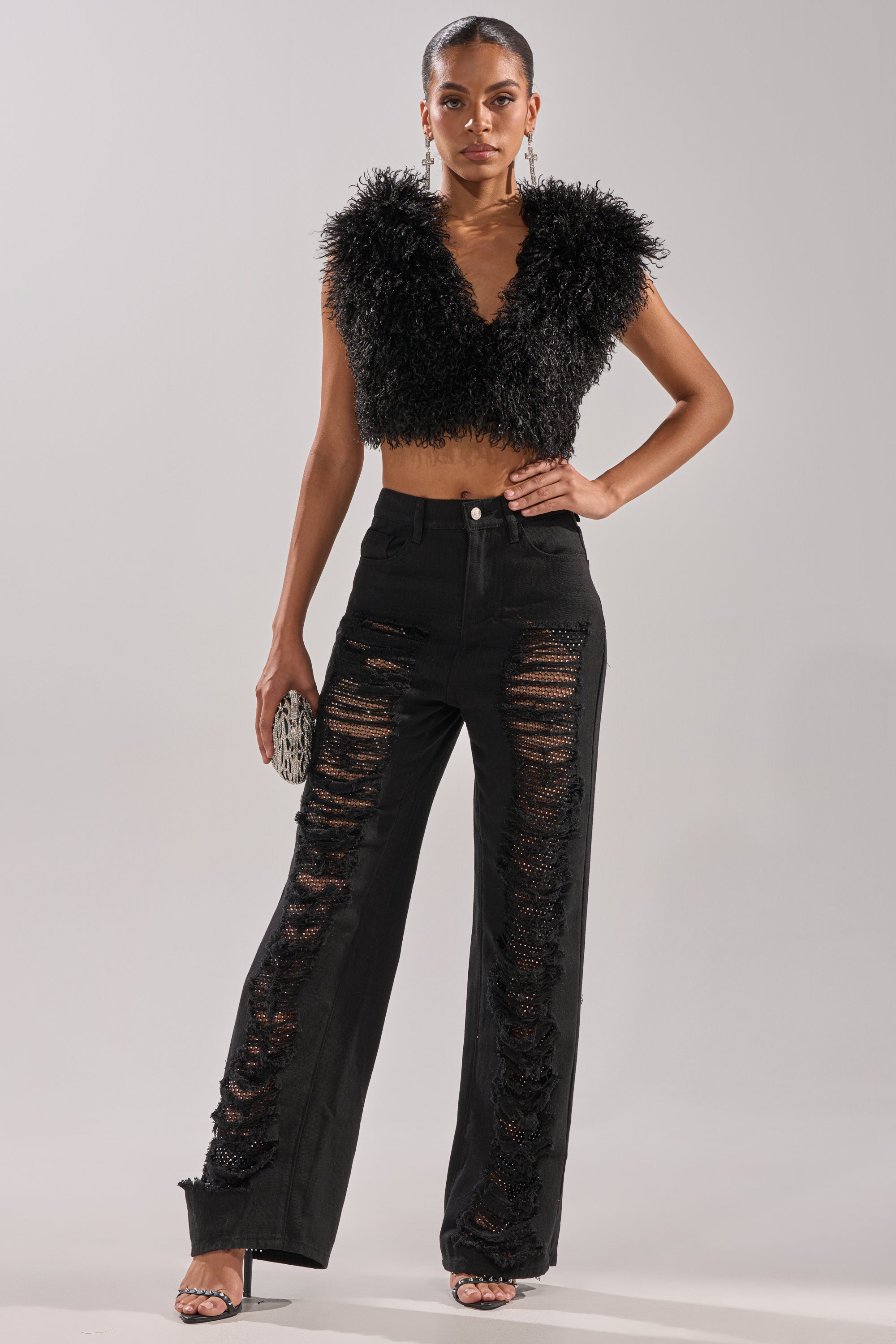 A woman exudes confidence in CLARA DISTRESSED DENIM high-rise jeans with large, ripped mesh panels over a rhinestone fishnet overlay, paired with a black fuzzy crop top, black heeled sandals, and a small silver clutch.