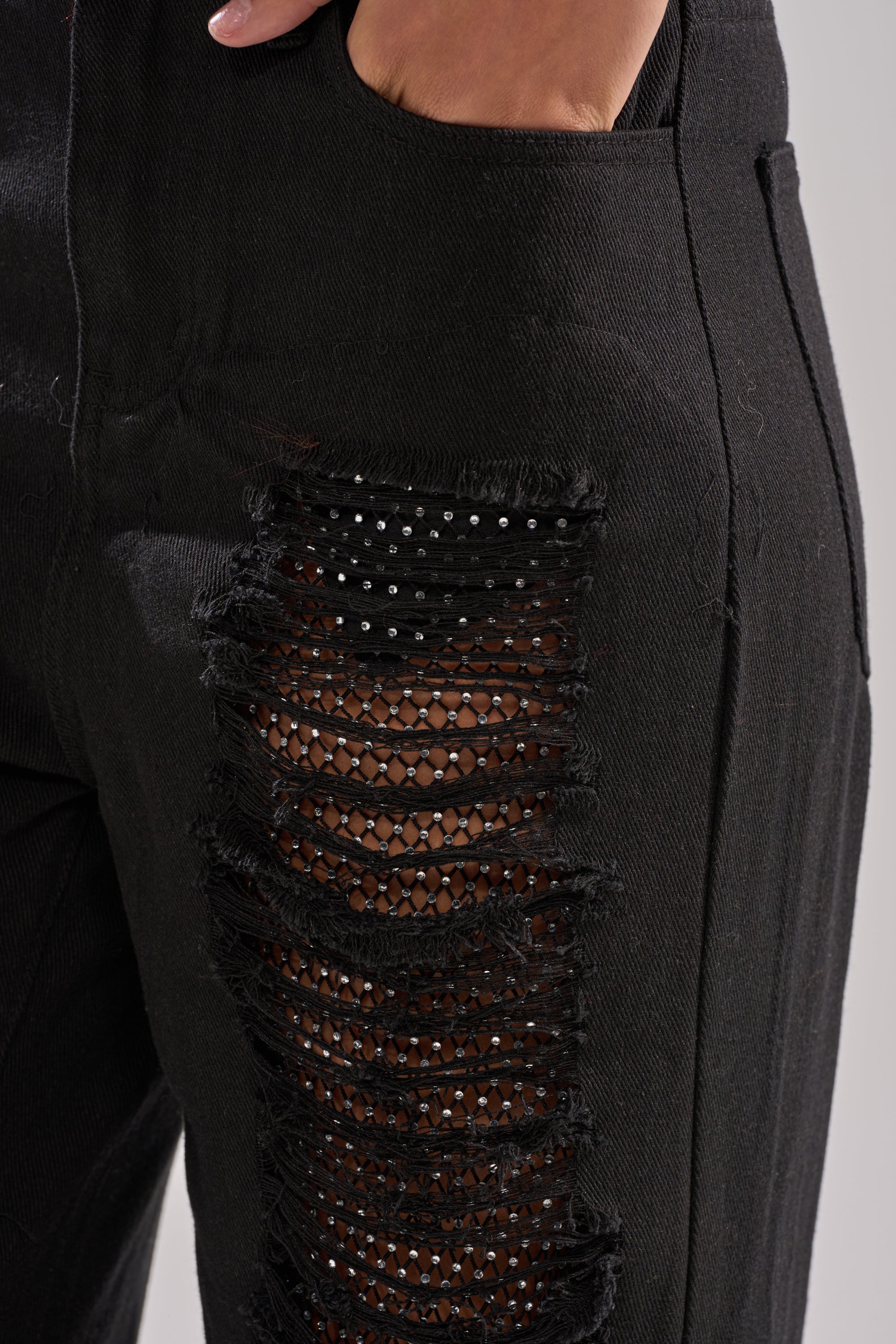 A close-up of a person wearing CLARA DISTRESSED DENIM black jeans with a hand in the pocket. These high-rise jeans showcase horizontal rips, decorative black mesh, and edgy small studs underneath.