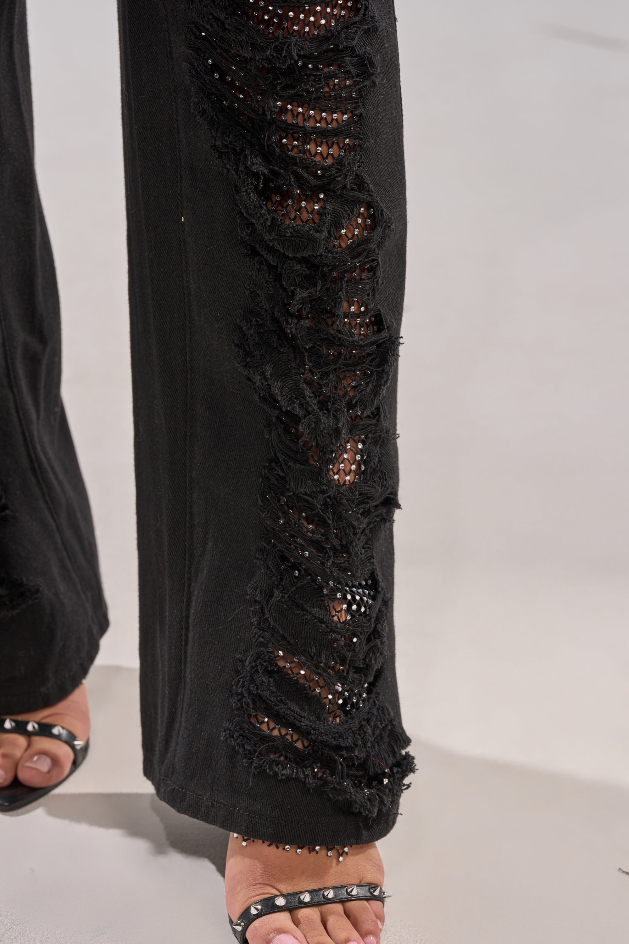 Close-up of a person wearing the CLARA DISTRESSED DENIM in black, featuring ripped detailing along the leg that reveals skin. The look is paired with black open-toe sandals adorned with small metallic studs.