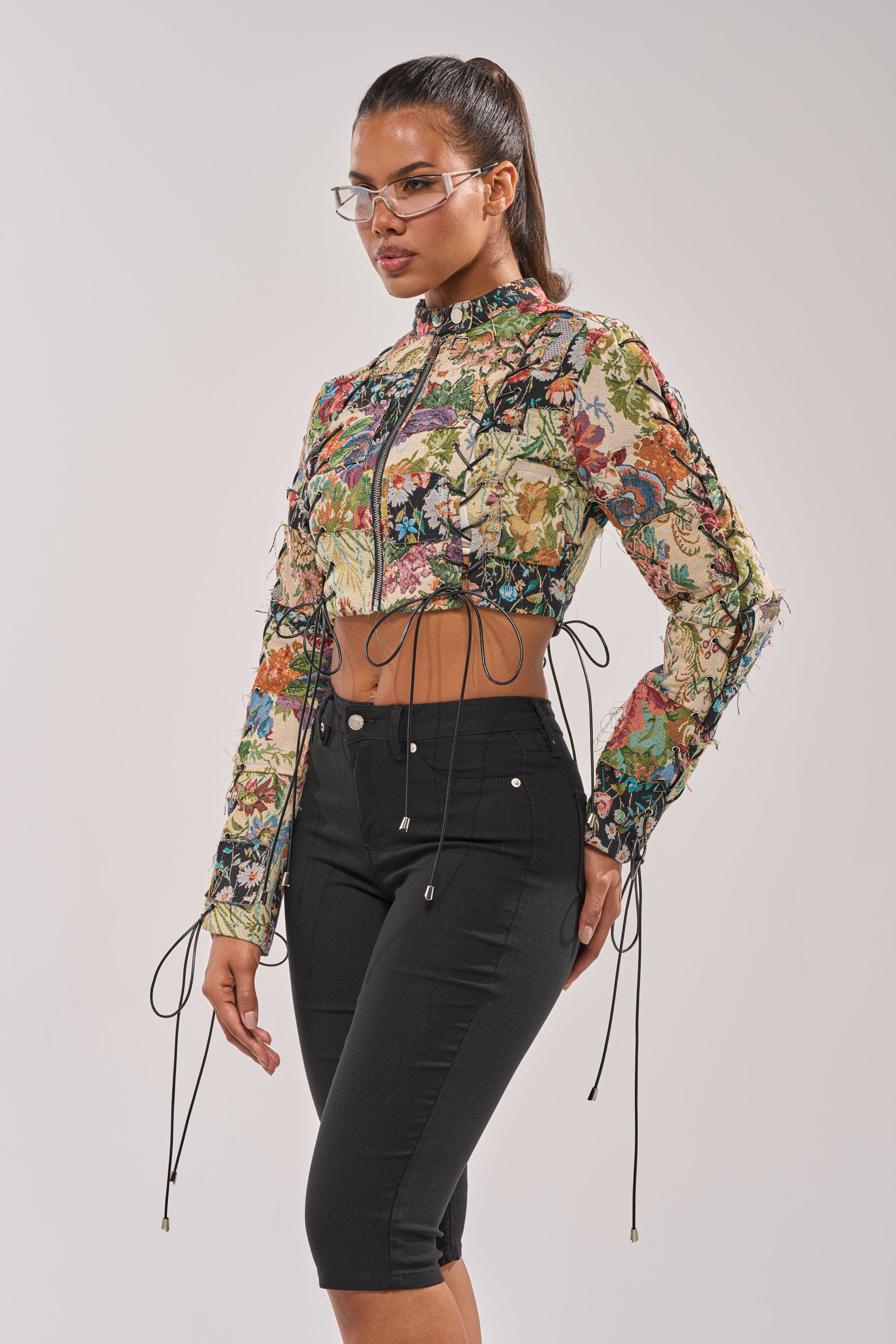 A woman with a high ponytail and glasses wears the TELL ME ABOUT IT PATCHWORK MOTO jacket and high-waisted black pants, standing against a plain light background. 0