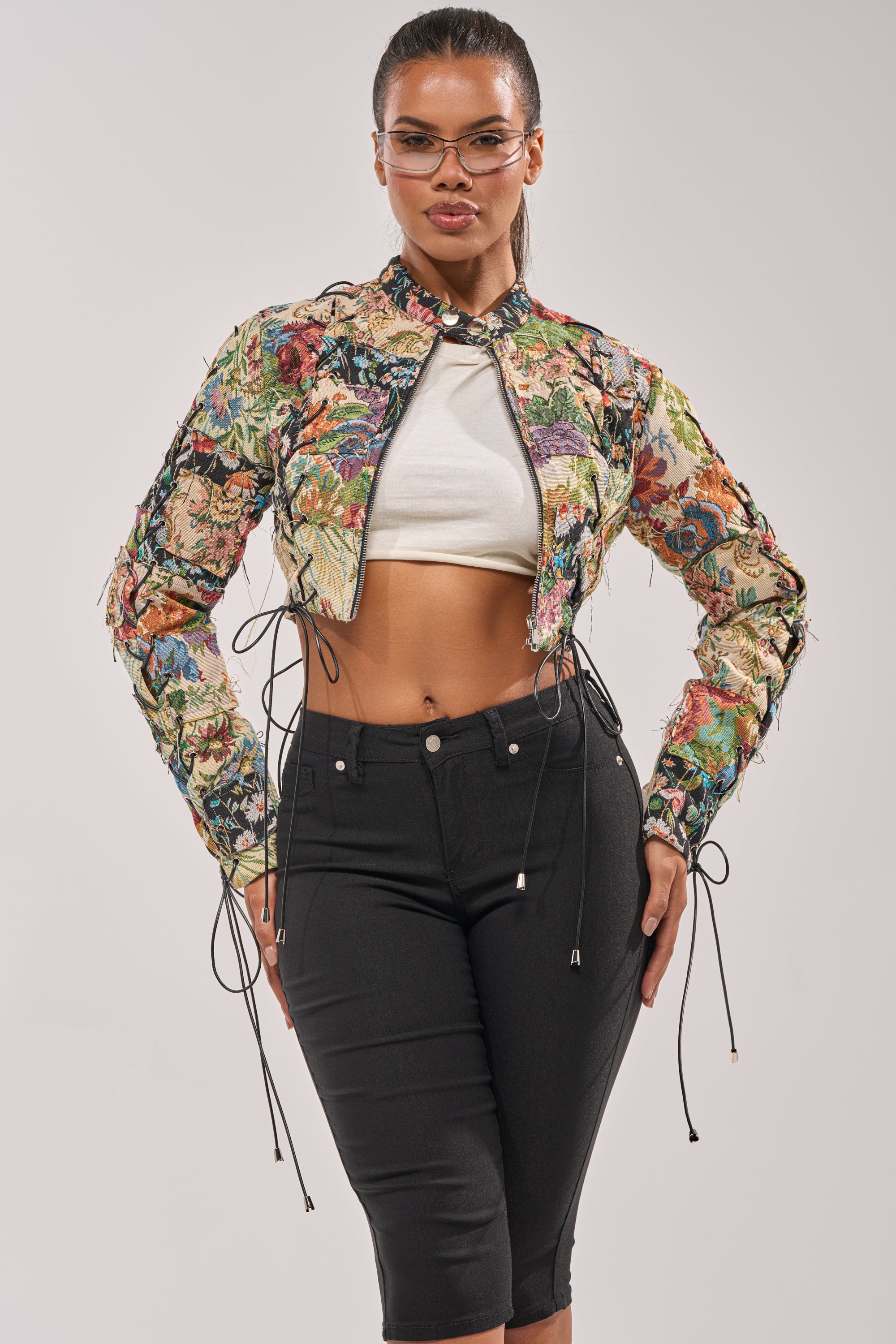 A woman poses confidently in the TELL ME ABOUT IT PATCHWORK MOTO jacket, styled with glasses, a white crop top, and high-waisted black pants against a plain light background. 2