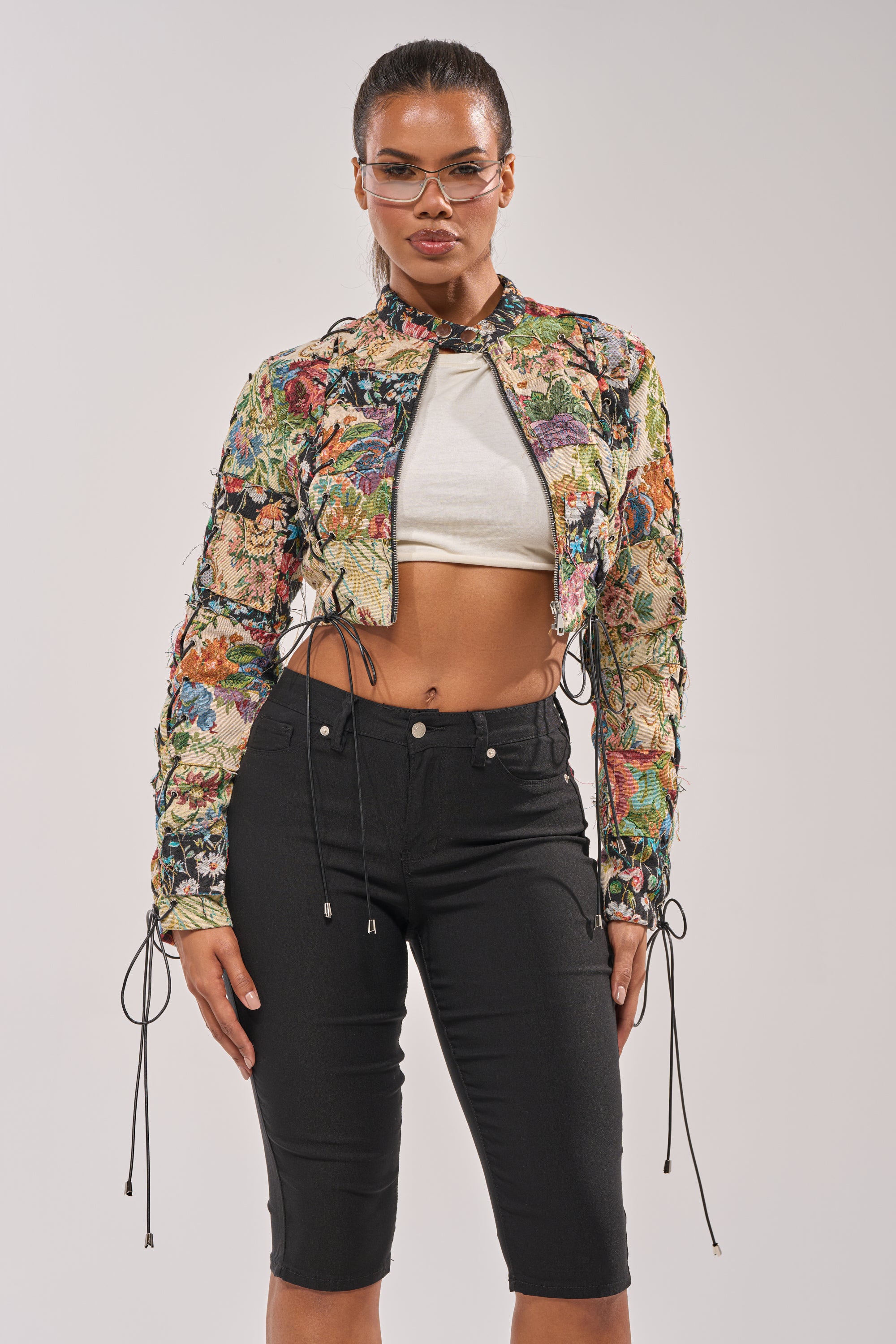 A woman poses confidently in the TELL ME ABOUT IT PATCHWORK MOTO jacket, paired with a white crop top, black fitted knee-length pants, and clear glasses against a plain background. 3