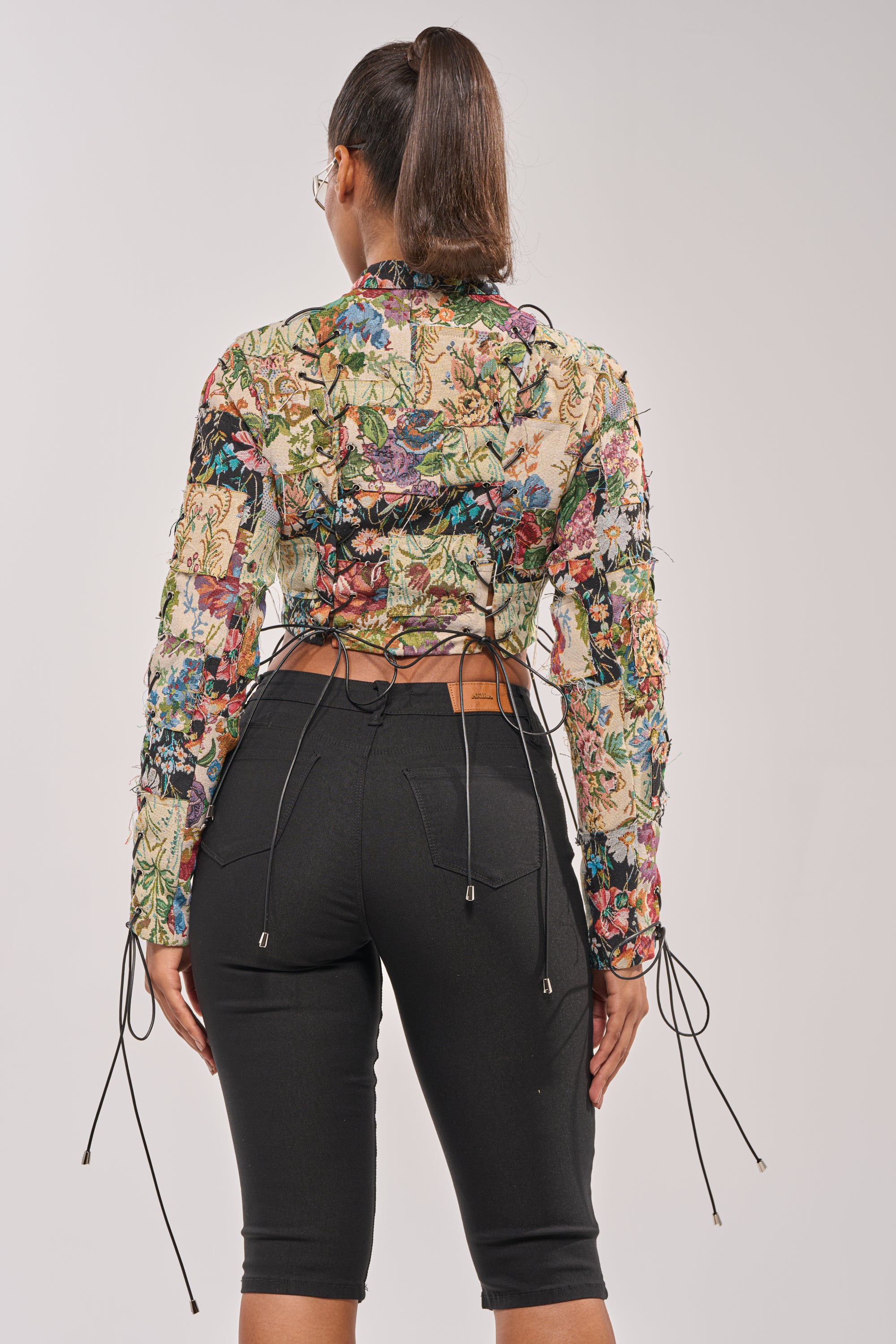 A woman with her hair in a ponytail stands with her back to the camera wearing the TELL ME ABOUT IT PATCHWORK MOTO, a colorful patchwork moto-style top, paired with fitted black knee-length pants. 5