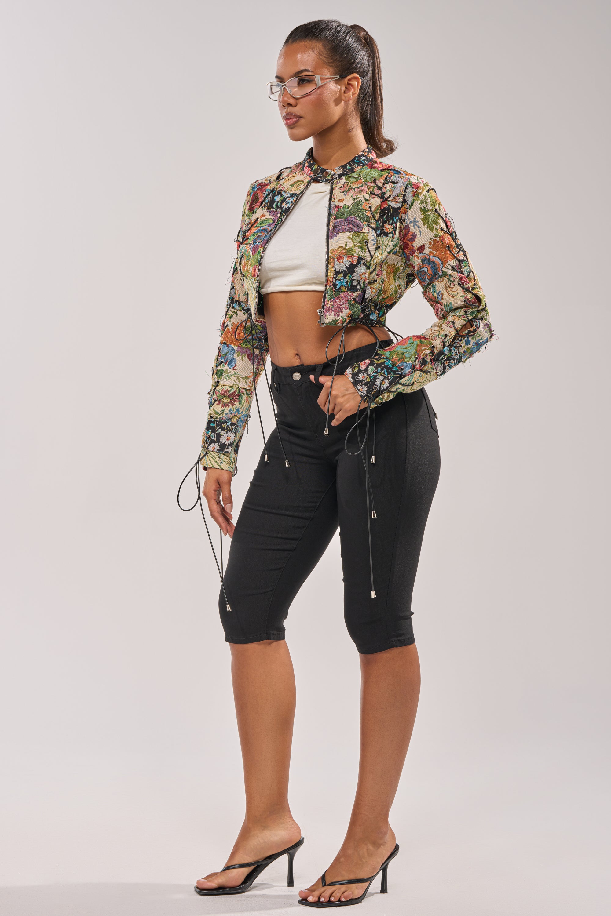 Woman standing sideways wearing the TELL ME ABOUT IT PATCHWORK MOTO jacket, a white crop top, black high-waisted knee-length shorts, black heeled sandals, and glasses against a plain background. 6