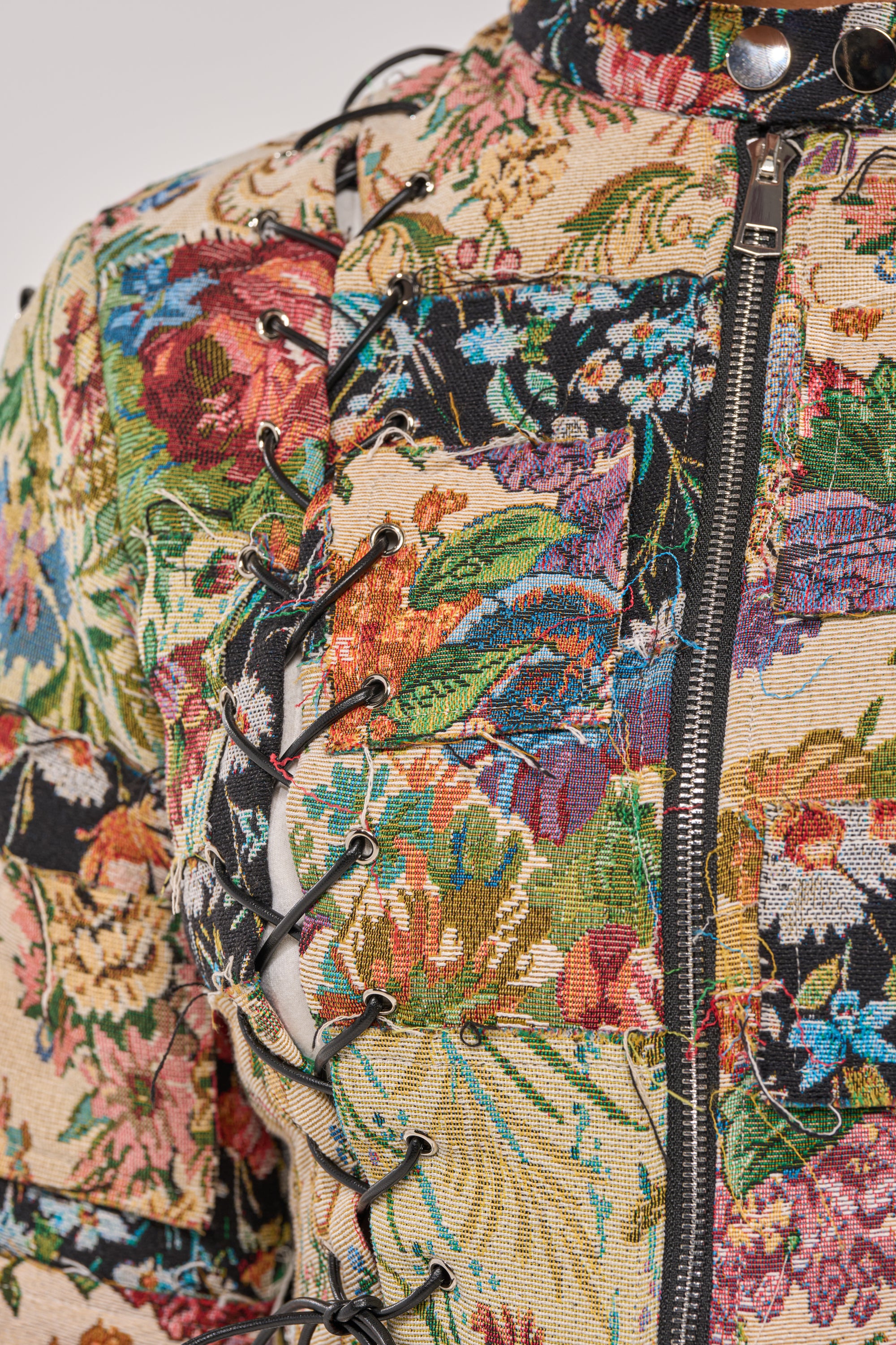 Close-up of the TELL ME ABOUT IT PATCHWORK MOTO jacket, featuring colorful brocade patchwork, a visible zipper, and black leather lacing along the seams. 7