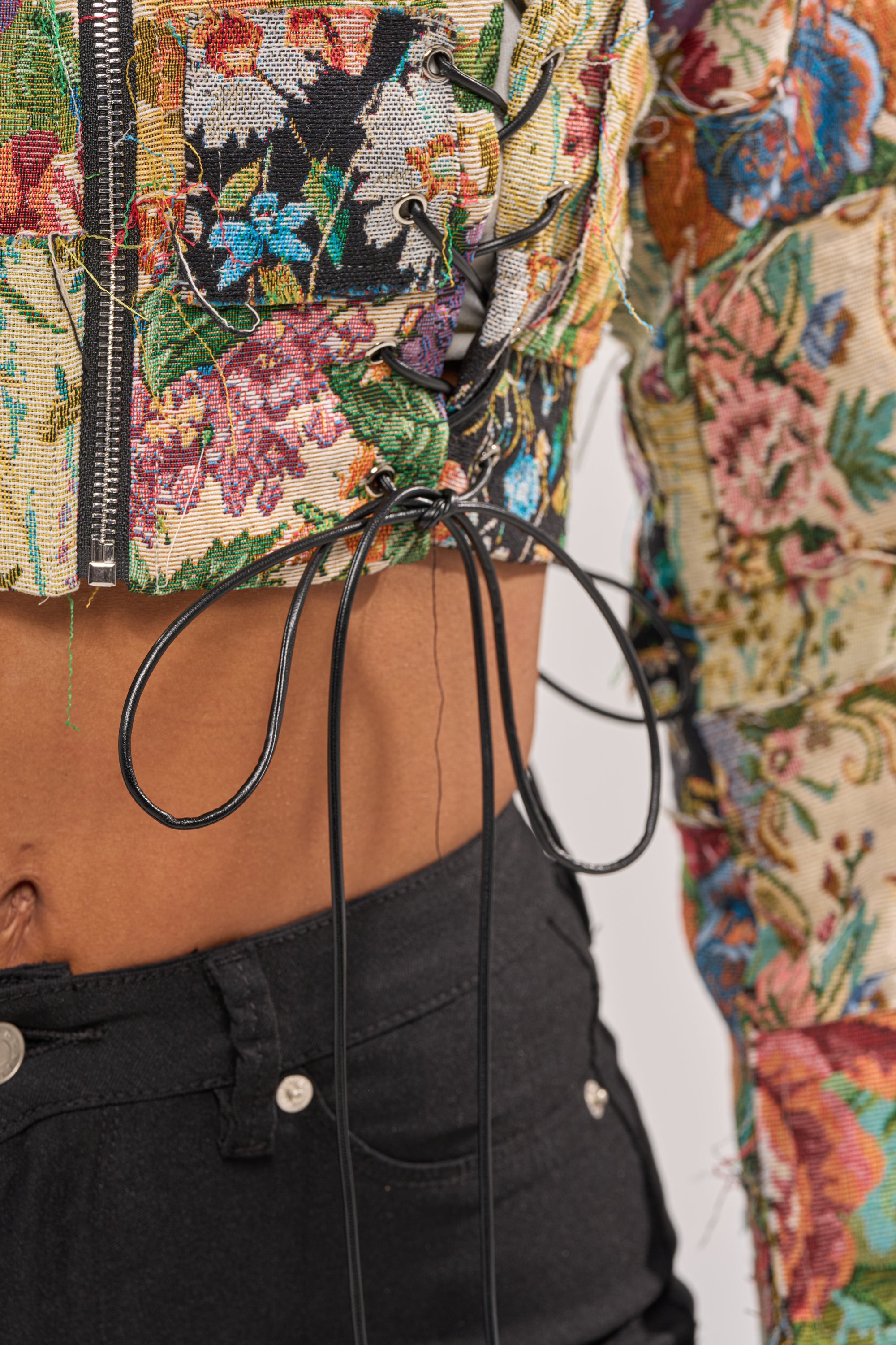 A close-up of a person wearing high-waisted black jeans and the TELL ME ABOUT IT PATCHWORK MOTO, a colorful floral brocade cropped jacket with exposed stitching, black cord lacing at the waist, and a midriff-baring moto style. 8