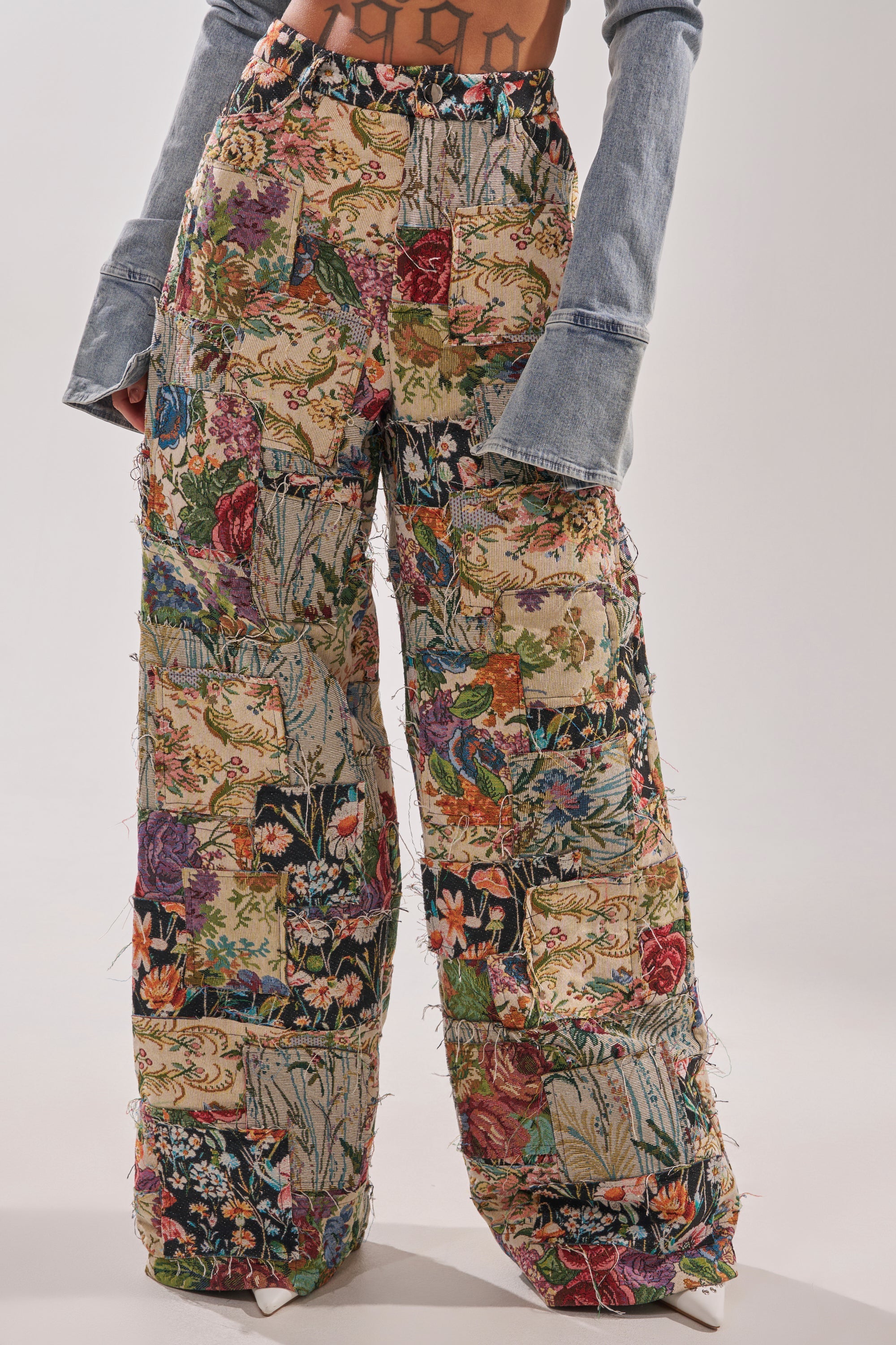 Only the lower torso and legs are visible, wearing the OUT OF MY MIND TAPESTRY PANT—high-waisted, wide-leg pants with a colorful floral pattern—paired with a denim crop top with long flared sleeves. 0