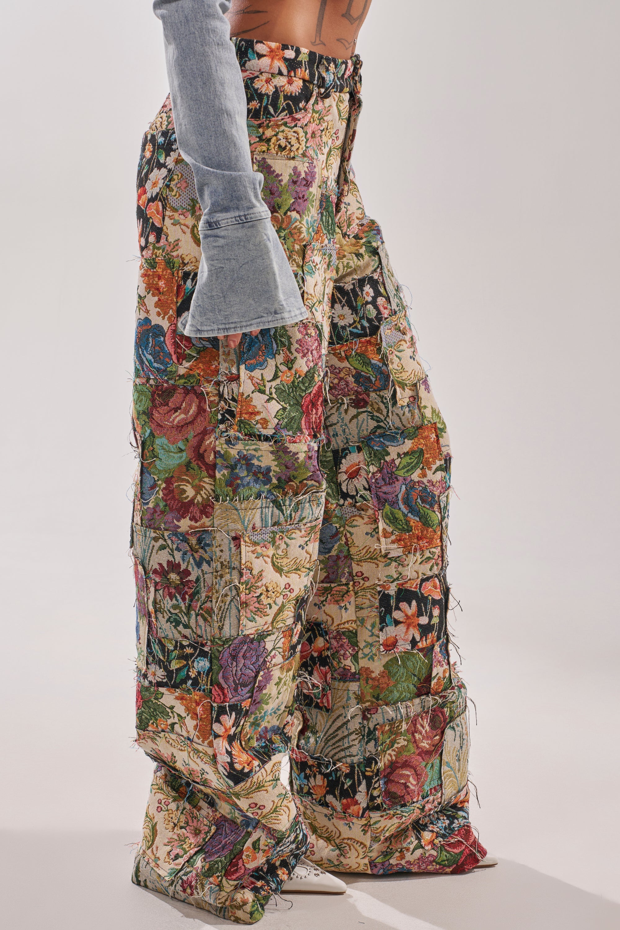 Someone wears the OUT OF MY MIND TAPESTRY PANT, featuring a wide-leg fit and vibrant patchwork floral design, paired with a light blue flared denim sleeve top, standing against a plain background. 2