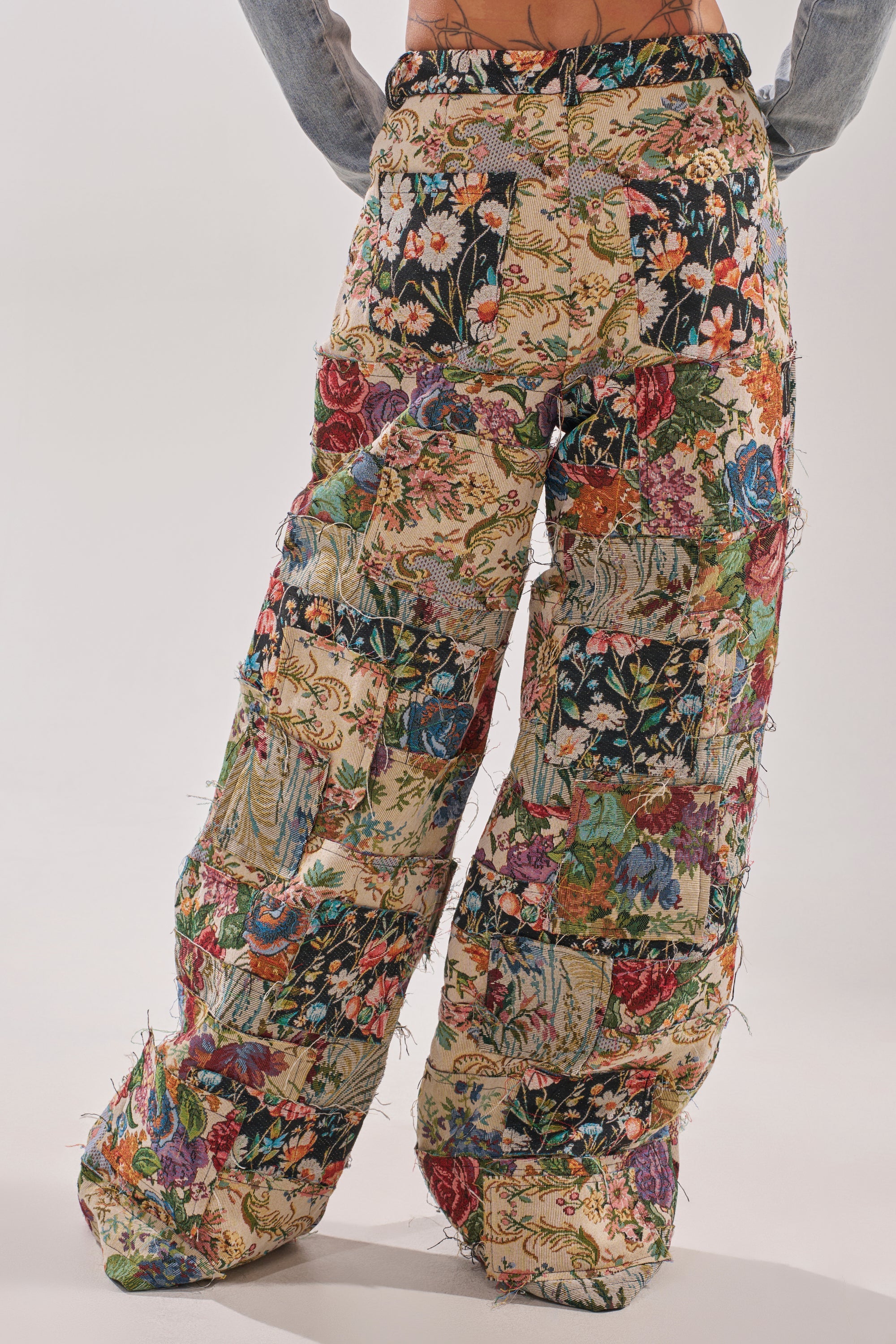 A person wearing OUT OF MY MIND TAPESTRY PANT—vintage-inspired wide-leg patchwork pants with colorful floral and nature patterns—stands against a plain light background. 3