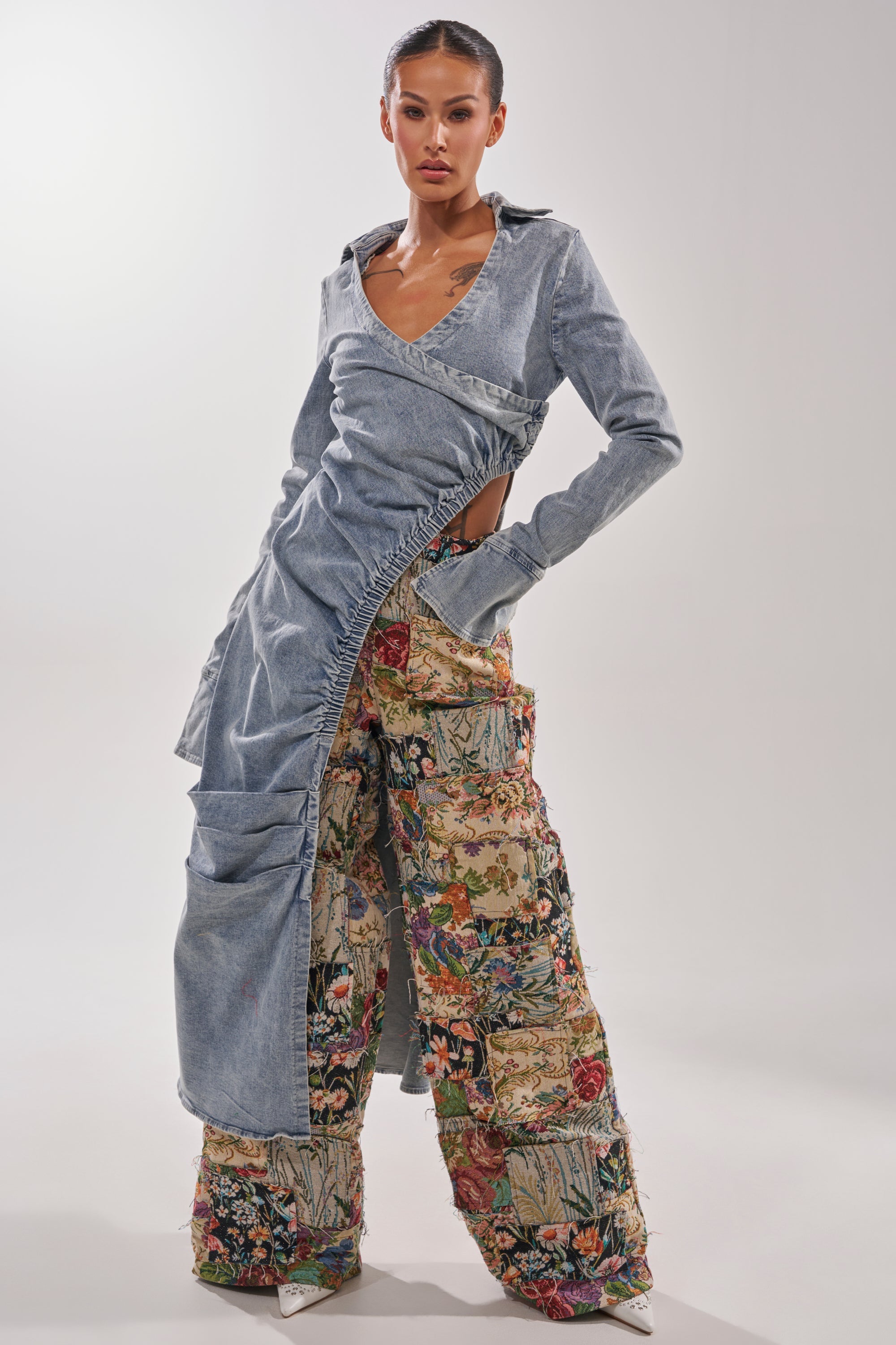 A woman poses against a plain, light background wearing the OUT OF MY MIND TAPESTRY PANT—wide-leg pants with colorful floral and patchwork designs—paired with a light blue, asymmetrical denim dress featuring an open side. 4