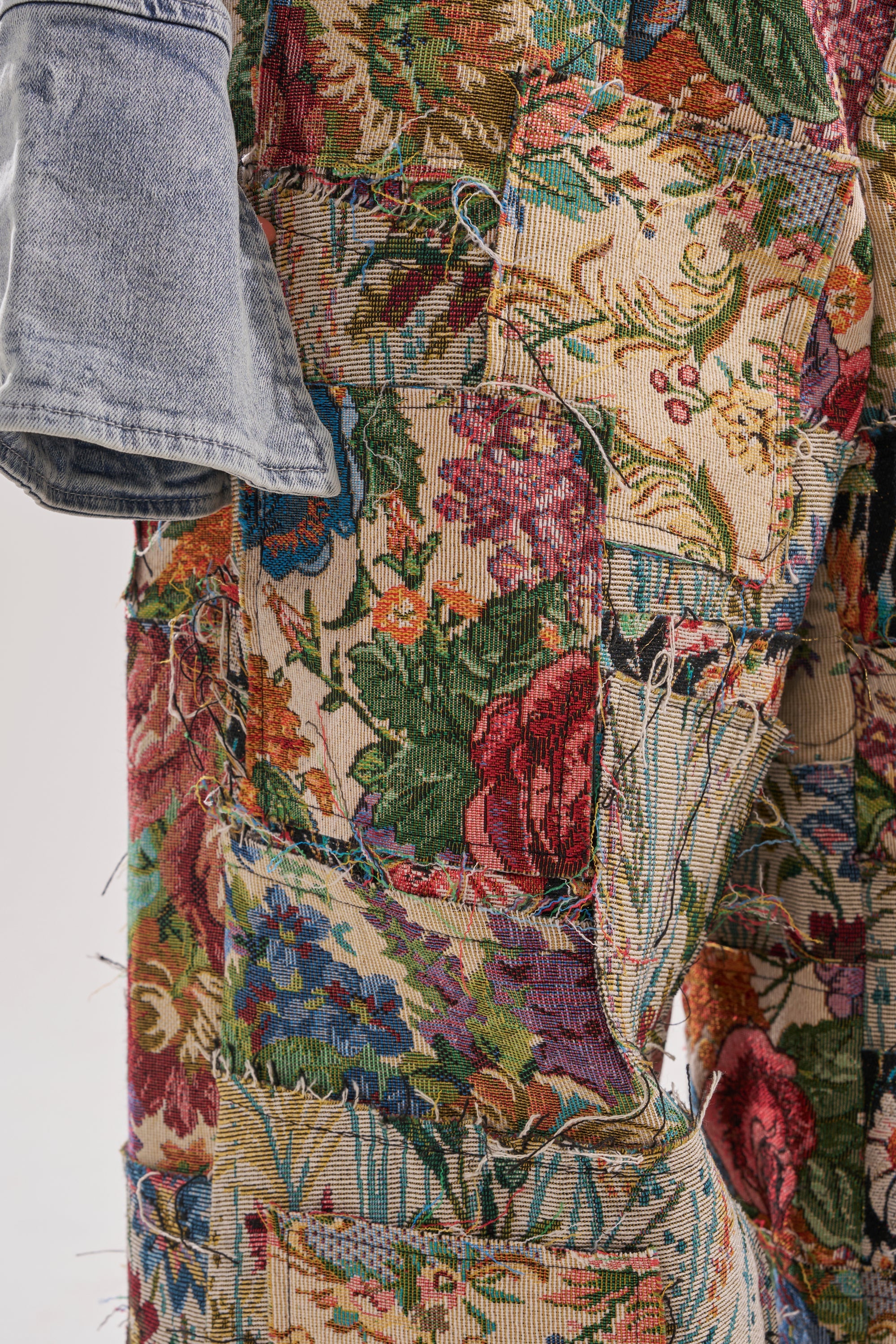 Close-up of the OUT OF MY MIND TAPESTRY PANT, featuring colorful floral-patterned fabric with visible stitching and frayed edges, paired with a blue denim jacket sleeve. 6