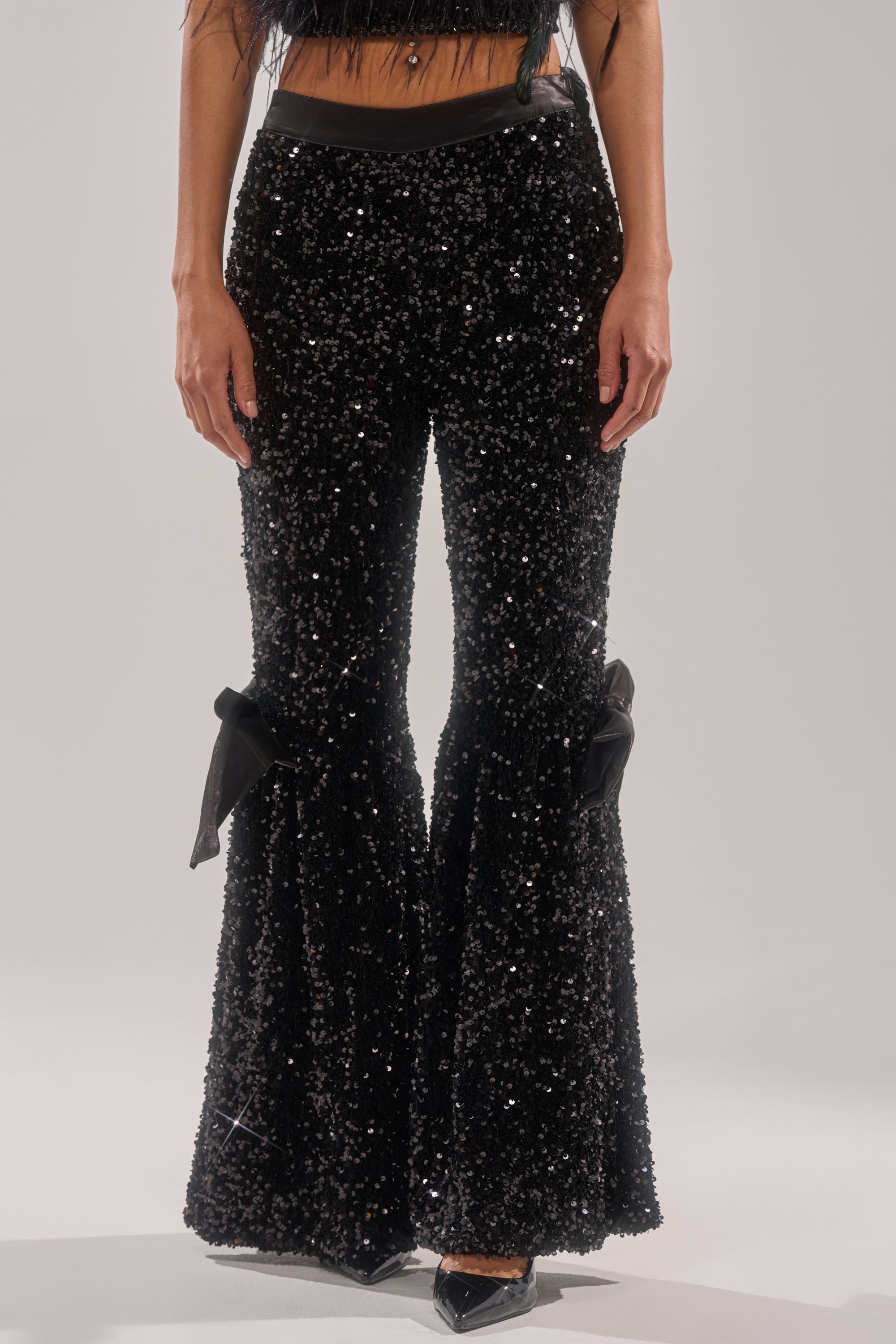 A person models the SELENA SPARKLING WIDE LEG PANT, featuring large black bows at the knees, paired with a black top and pointed-toe shoes against a plain light background. 1