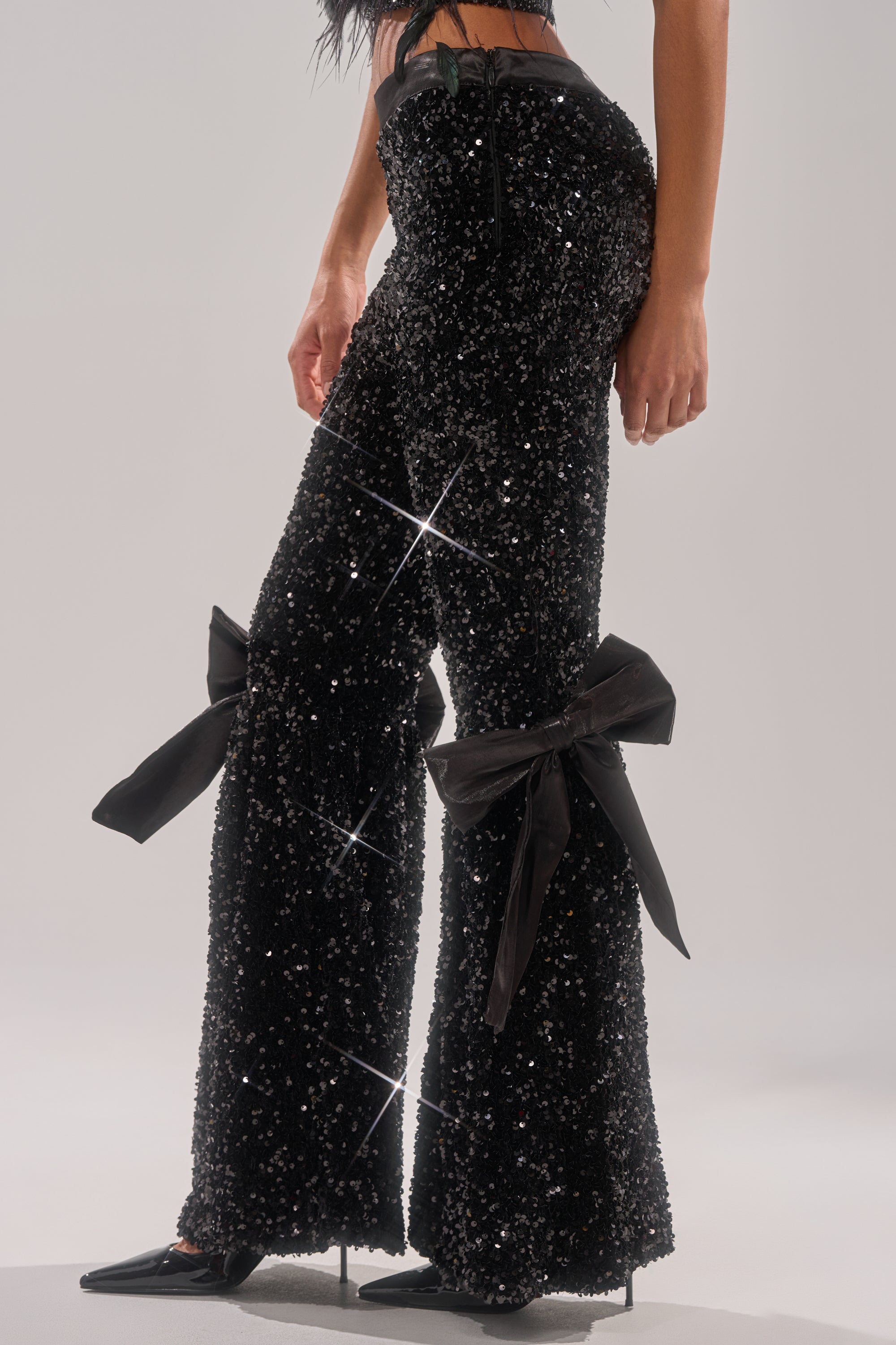 A person wears the SELENA SPARKLING WIDE LEG PANT, featuring large satin bows on the legs, paired with black pointed heels. The photo is cropped at the waist and ankles to highlight the pants’ shimmering detail. 2