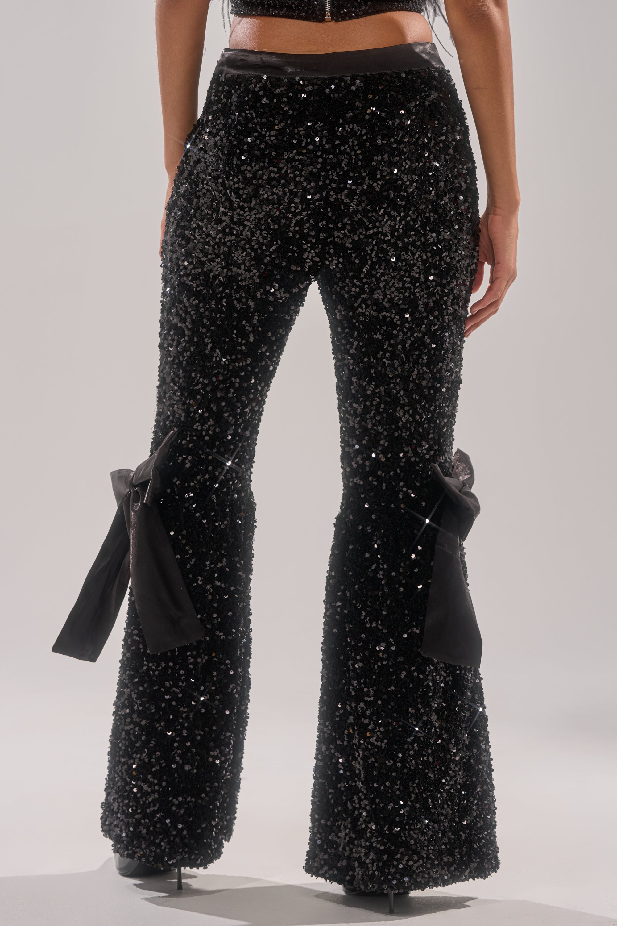A person shown from the waist down wears SELENA SPARKLING WIDE LEG PANT with large black bows above the knees and black high-heeled shoes. 3