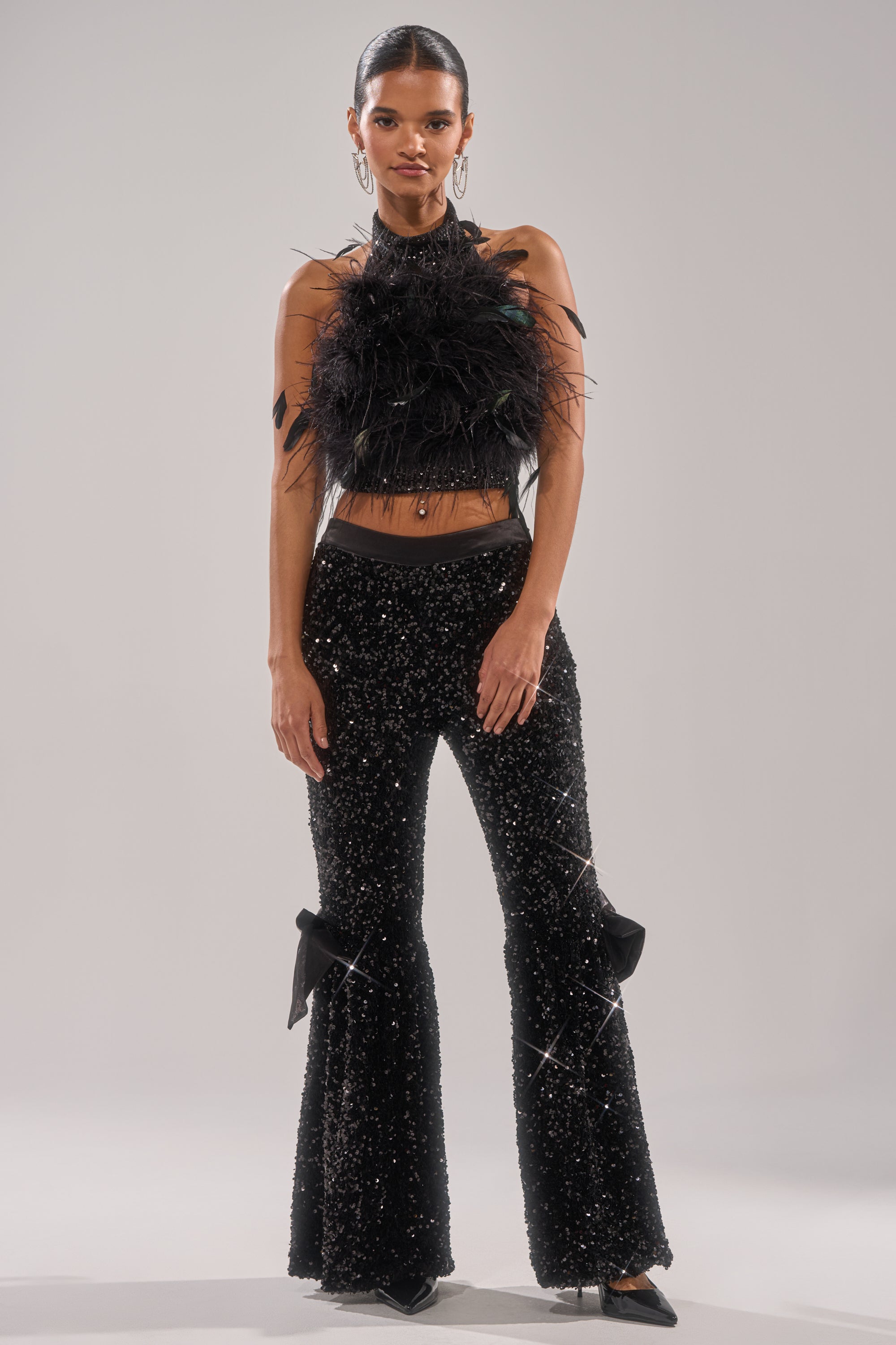 A woman poses confidently in SELENA SPARKLING WIDE LEG PANT—sparkly wide-leg pants with bow details—paired with a sleeveless black feathered crop top, hoop earrings, and sleek hair against a plain background. 4