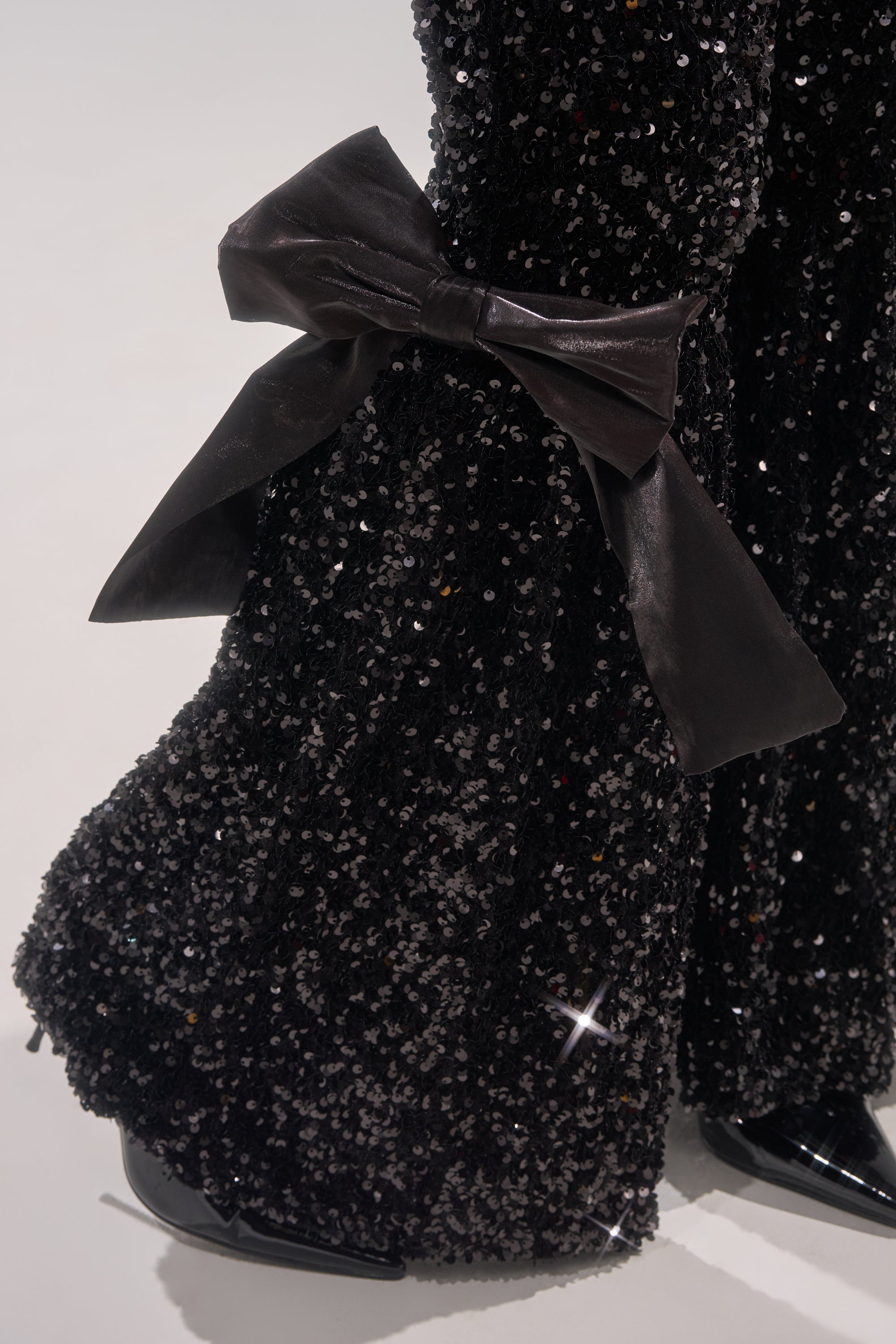 Close-up of a black sequined garment with a large black satin bow, paired with the SELENA SPARKLING WIDE LEG PANT and shiny black shoes against a plain light background. 6