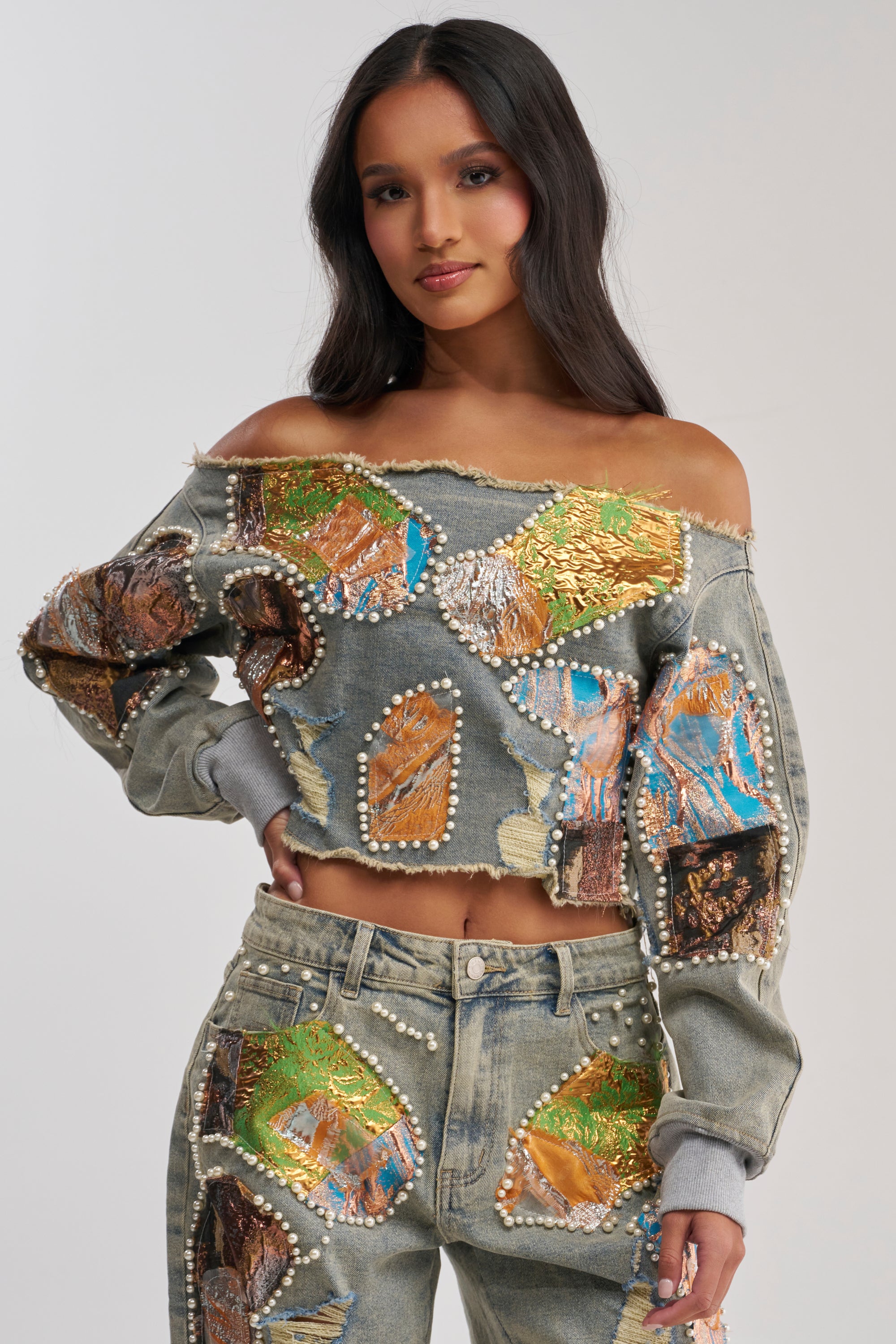 A woman with long, dark hair and a soft smile wears the PATCHED UP BROCADE DENIM SWEATSHIRT—a relaxed-fit, cropped denim piece with colorful patches, pearls, gold details, and frayed edges for a statement look.