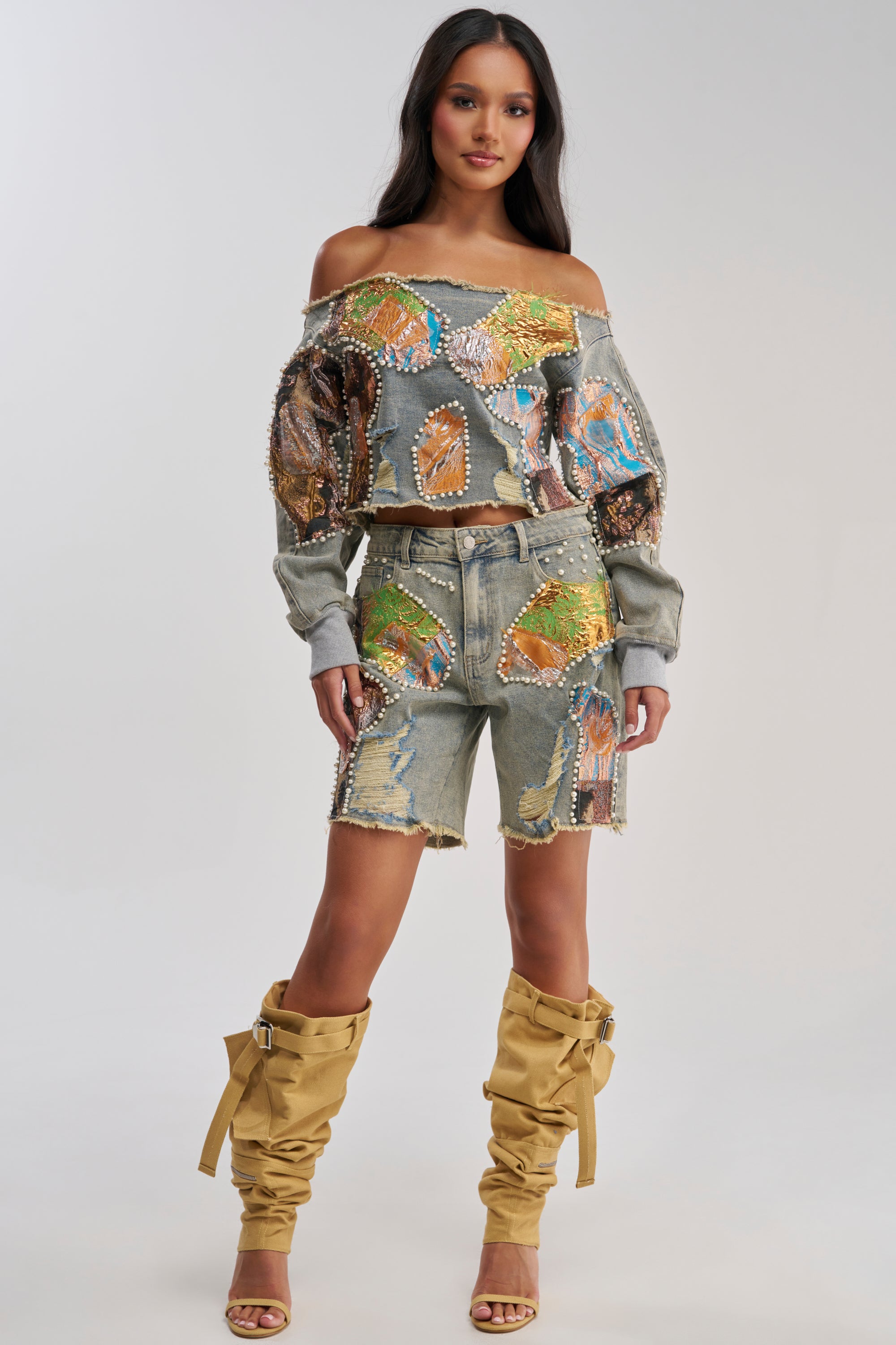 A woman wears the PATCHED UP LOW RISE BROCADE BERMUDA with an off-shoulder, embellished denim top. She pairs this chic look with tan slouchy knee-high boots and gold sandals, standing confidently against a light background.