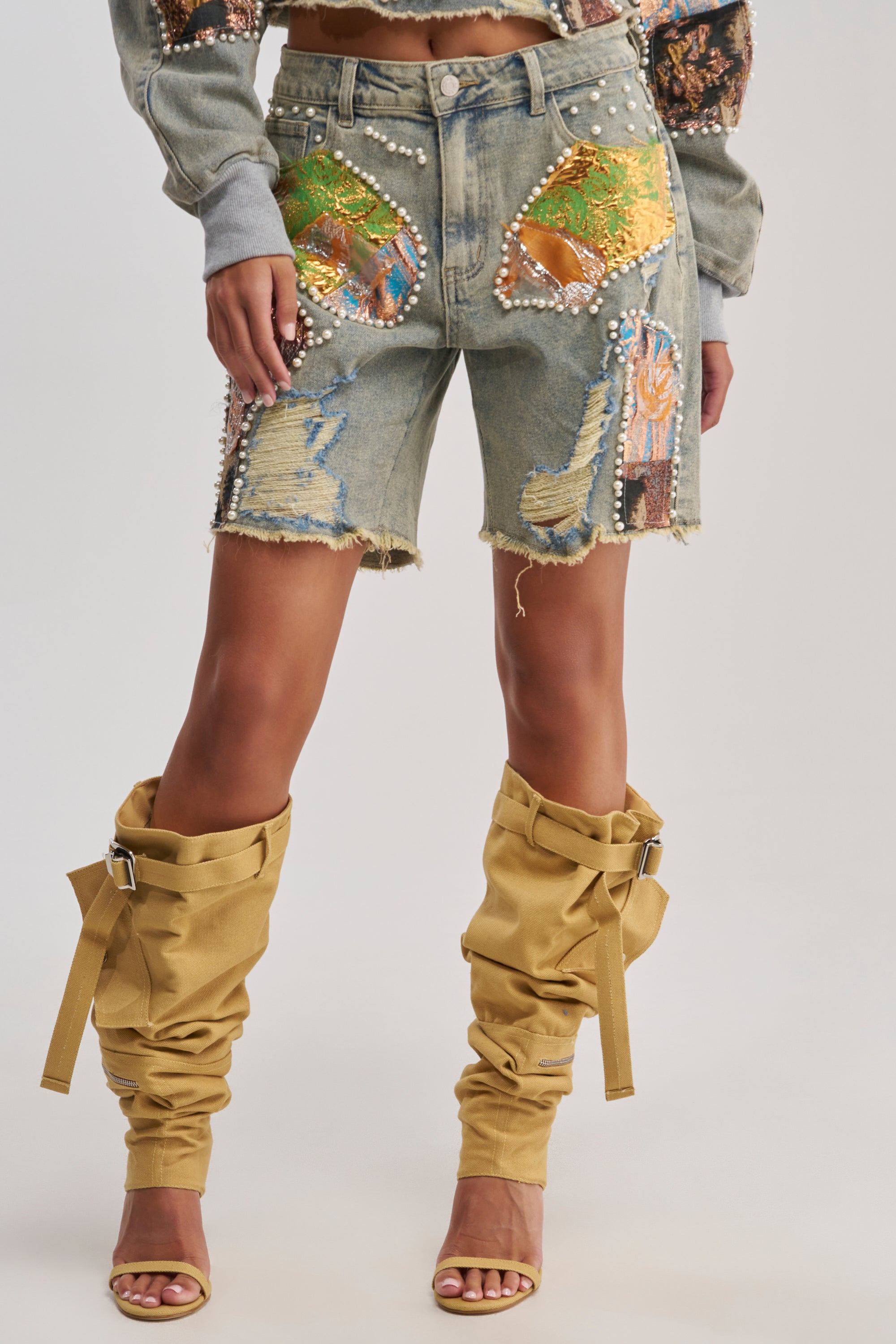Wearing the PATCHED UP LOW RISE BROCADE BERMUDA shorts with tan leg warmers and open-toe heels, a person creates a chic streetwear look as they rest their hands on their thighs.