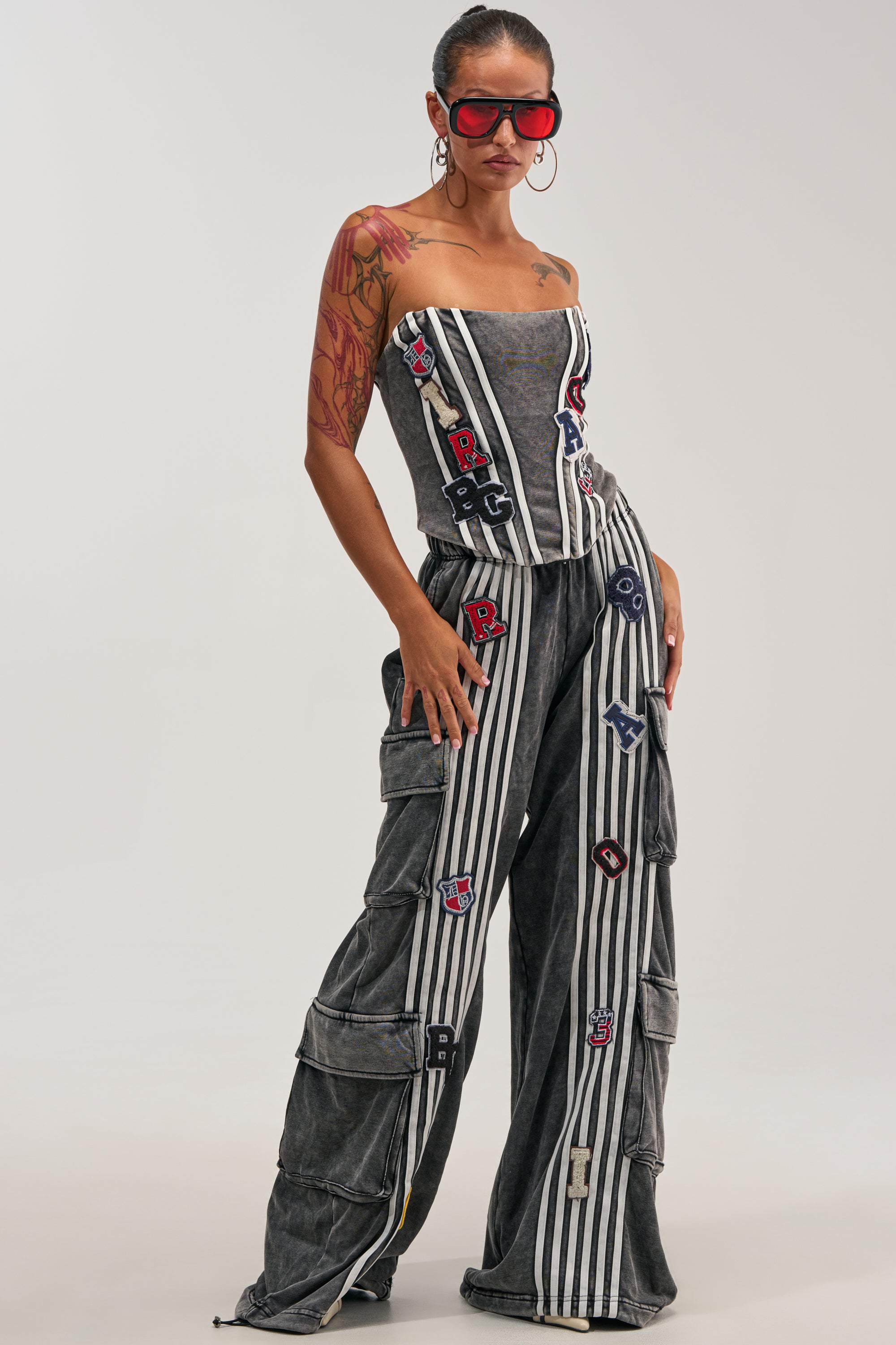 A woman stands confidently against a plain background wearing the ROCK AND ROLL WIDE LEG JOGGER, paired with a gray, striped, strapless top featuring colorful patches. She accessorizes her look with red sunglasses, hoop earrings, and shows off an arm tattoo.