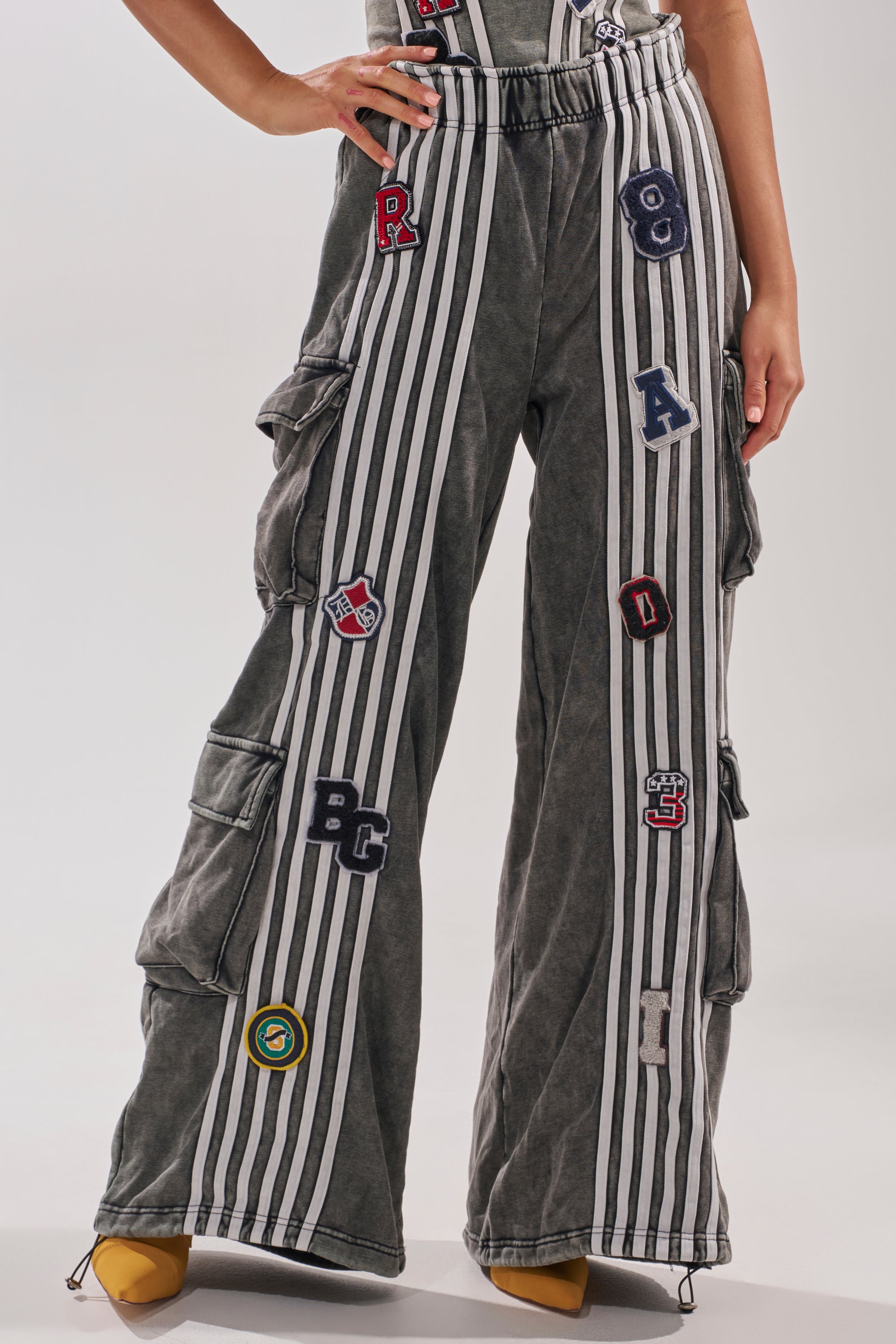 A person stands with one hand on their hip, wearing the ROCK AND ROLL WIDE LEG JOGGER featuring vertical white stripes, colorful patches, and yellow pointed shoes. The photo is cropped just below the chest. 2