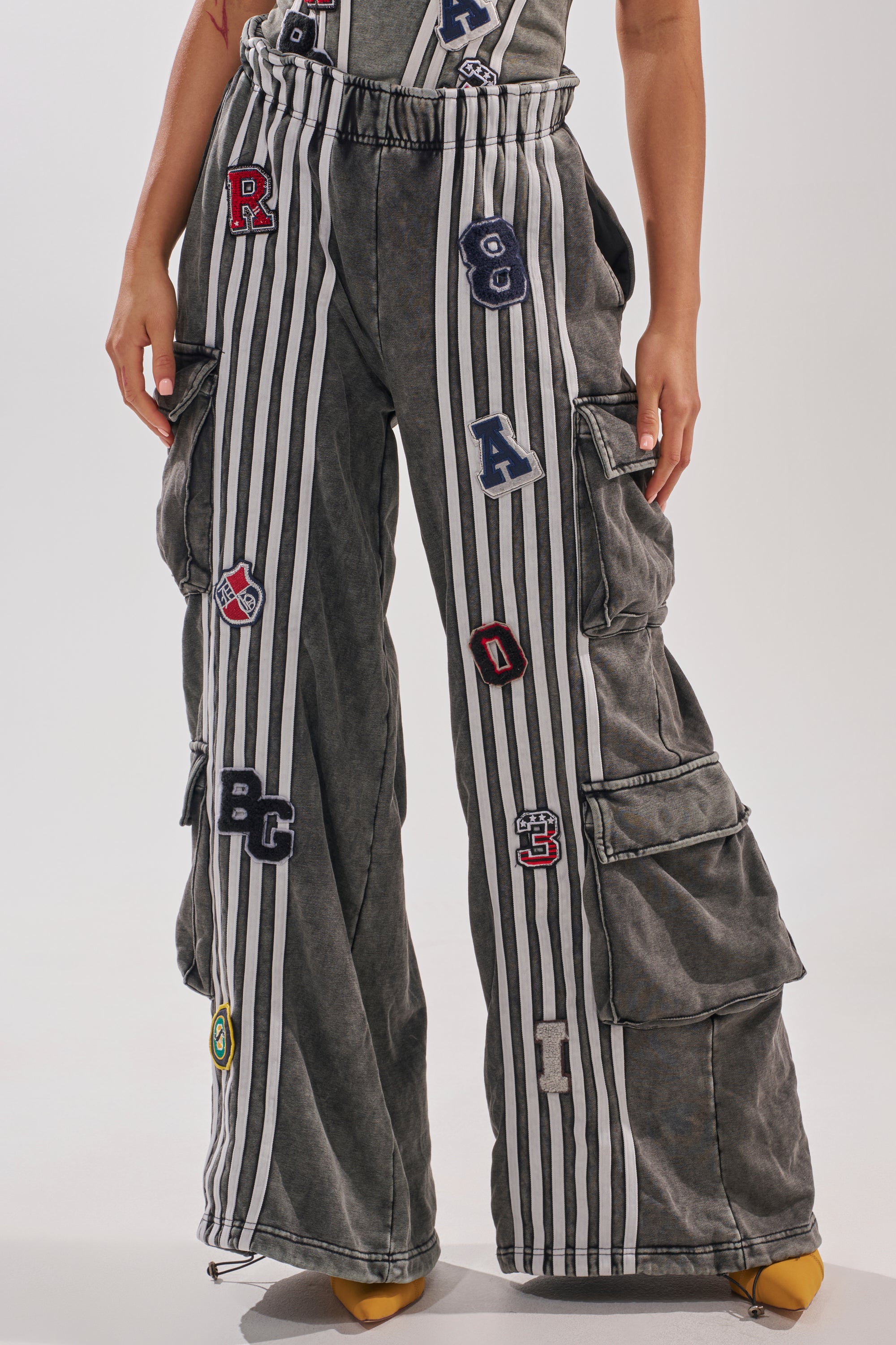 A person wears the AKIRA Label ROCK AND ROLL WIDE LEG JOGGER cargo pants featuring high waist, wide legs, vertical white stripes, colorful letter patches, and side pockets, paired with yellow pointed shoes. The upper body is partially out of frame. 0