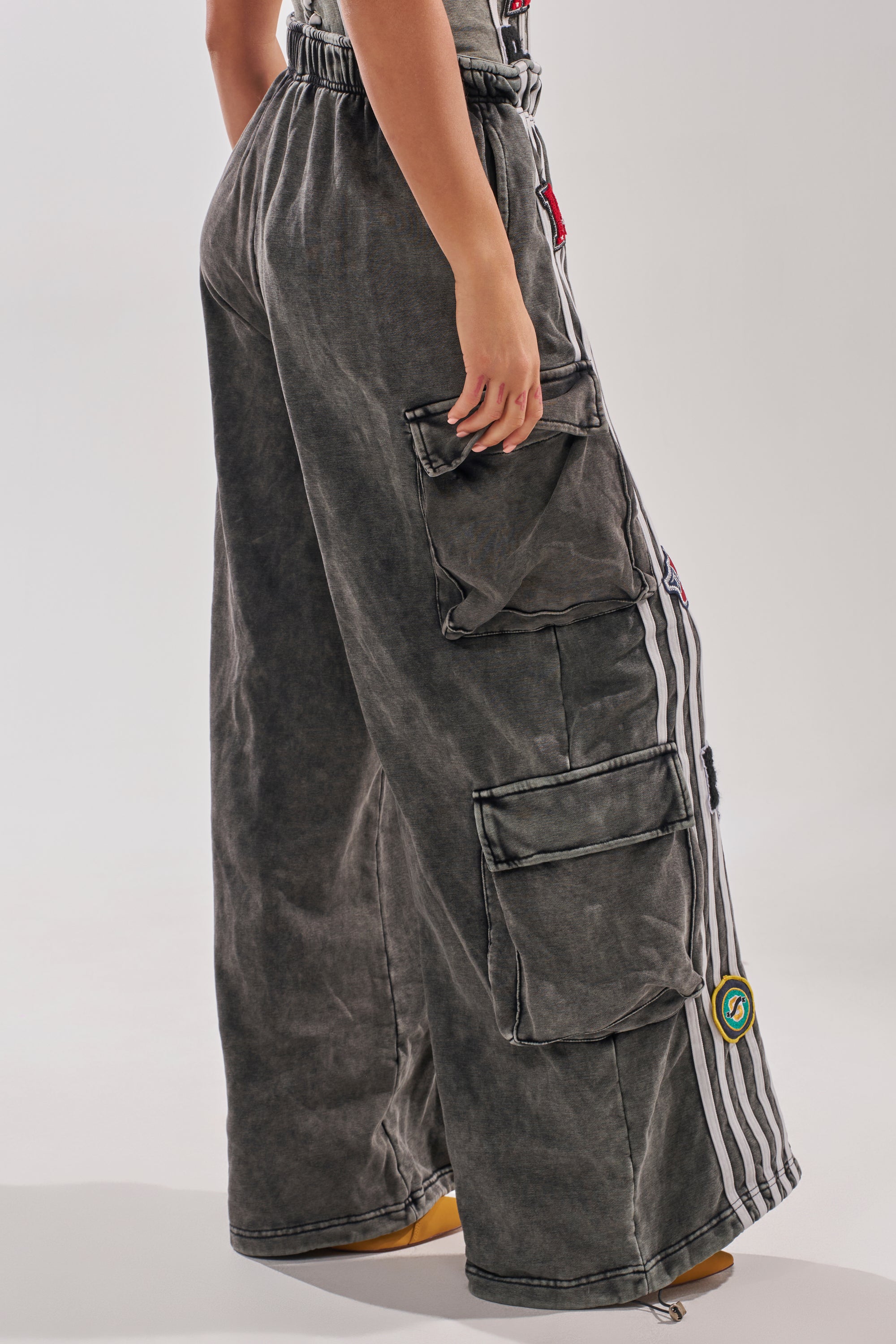 Wearing the ROCK AND ROLL WIDE LEG JOGGER with large pockets, white vertical stripes, and colorful patches, a person stands against a plain light background, showing only the lower half of their body. 3