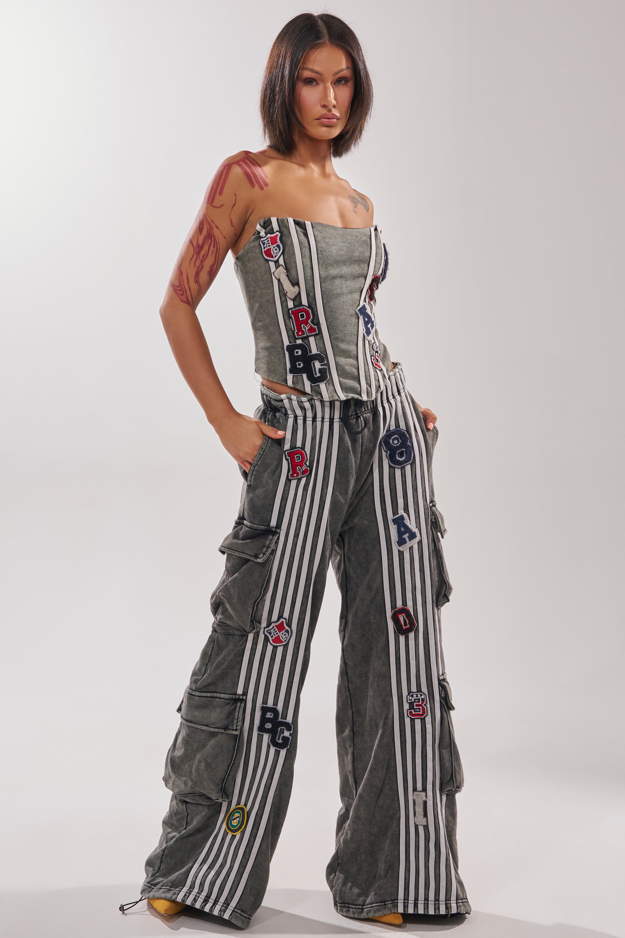 A woman with a bob haircut and arm tattoos poses confidently against a plain light background, wearing the ROCK AND ROLL WIDE LEG JOGGER—strapless, grey, with white stripes and colorful letter patches, paired with a matching corset top. 5