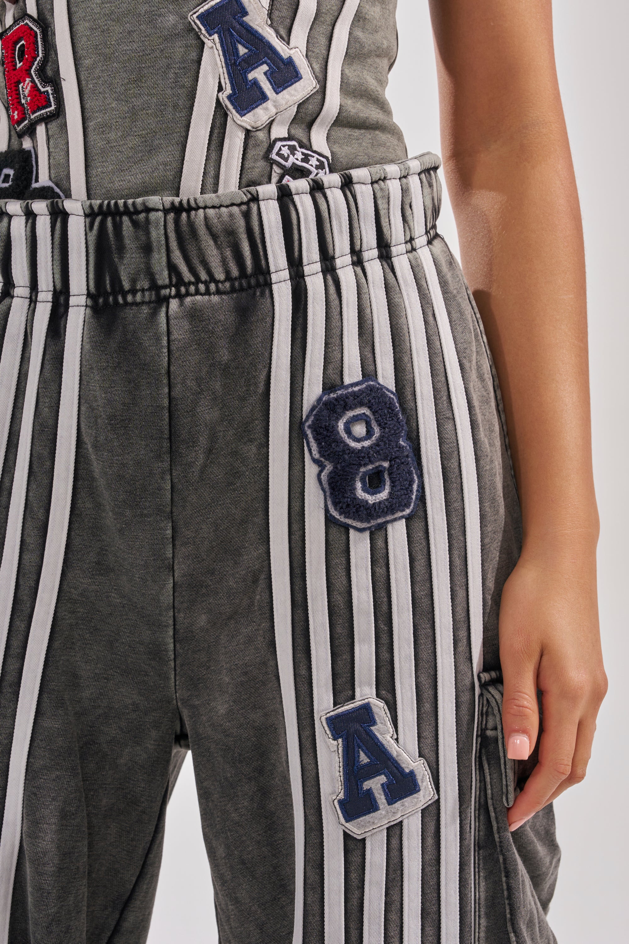 Someone wears the ROCK AND ROLL WIDE LEG JOGGER, a dark gray pant featuring white vertical stripes and blue-and-white patches with "8" and "A". Their right hand rests on their thigh. 6