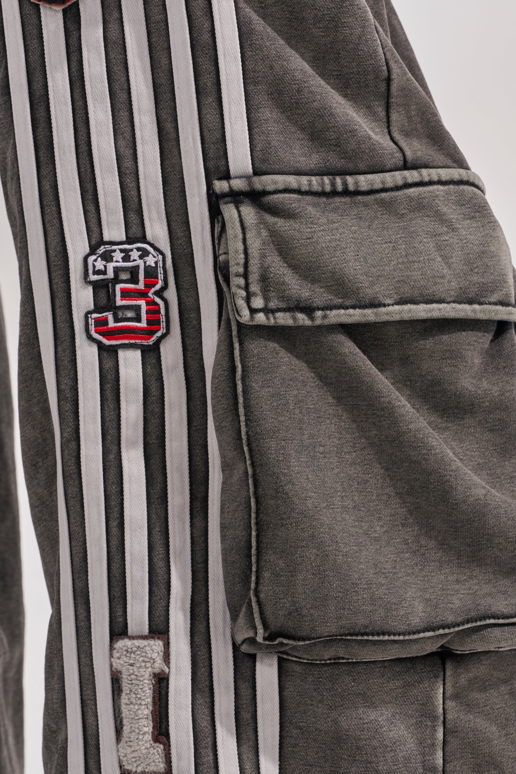 A close-up of a gray fabric sleeve with white vertical stripes, a large flap pocket, and embroidered patches, including a red and black number 5—signature details of the ROCK AND ROLL WIDE LEG JOGGER. A partially visible letter I is also seen. 7