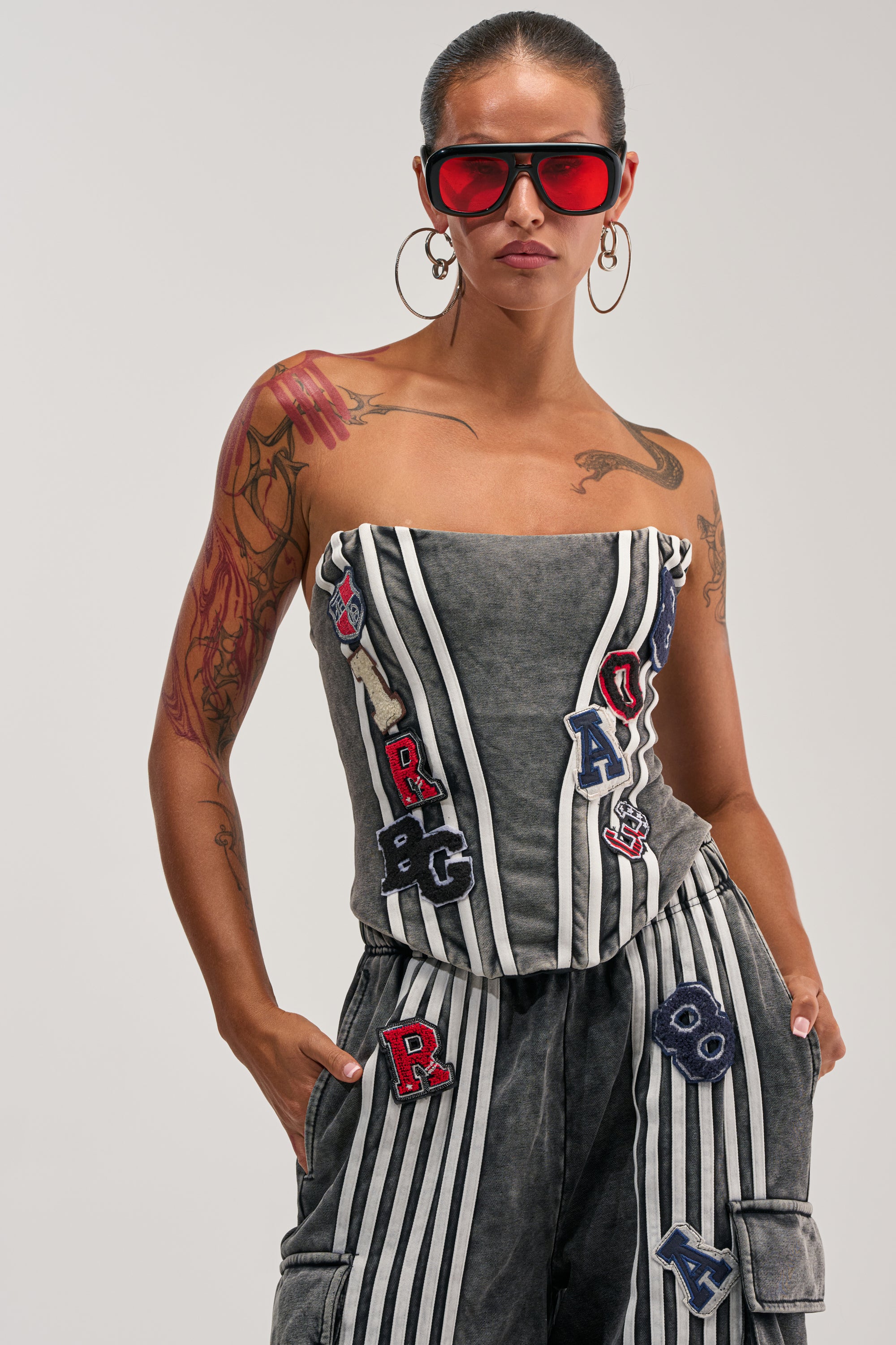 A woman with tattoos wears the ROCK AND ROLL CORSET, styled as a strapless, striped jumpsuit featuring letter patches and sporty-chic corset detailing. She completes her look with large hoop earrings and red sunglasses, standing confidently with her hands in her pockets against a plain background.
