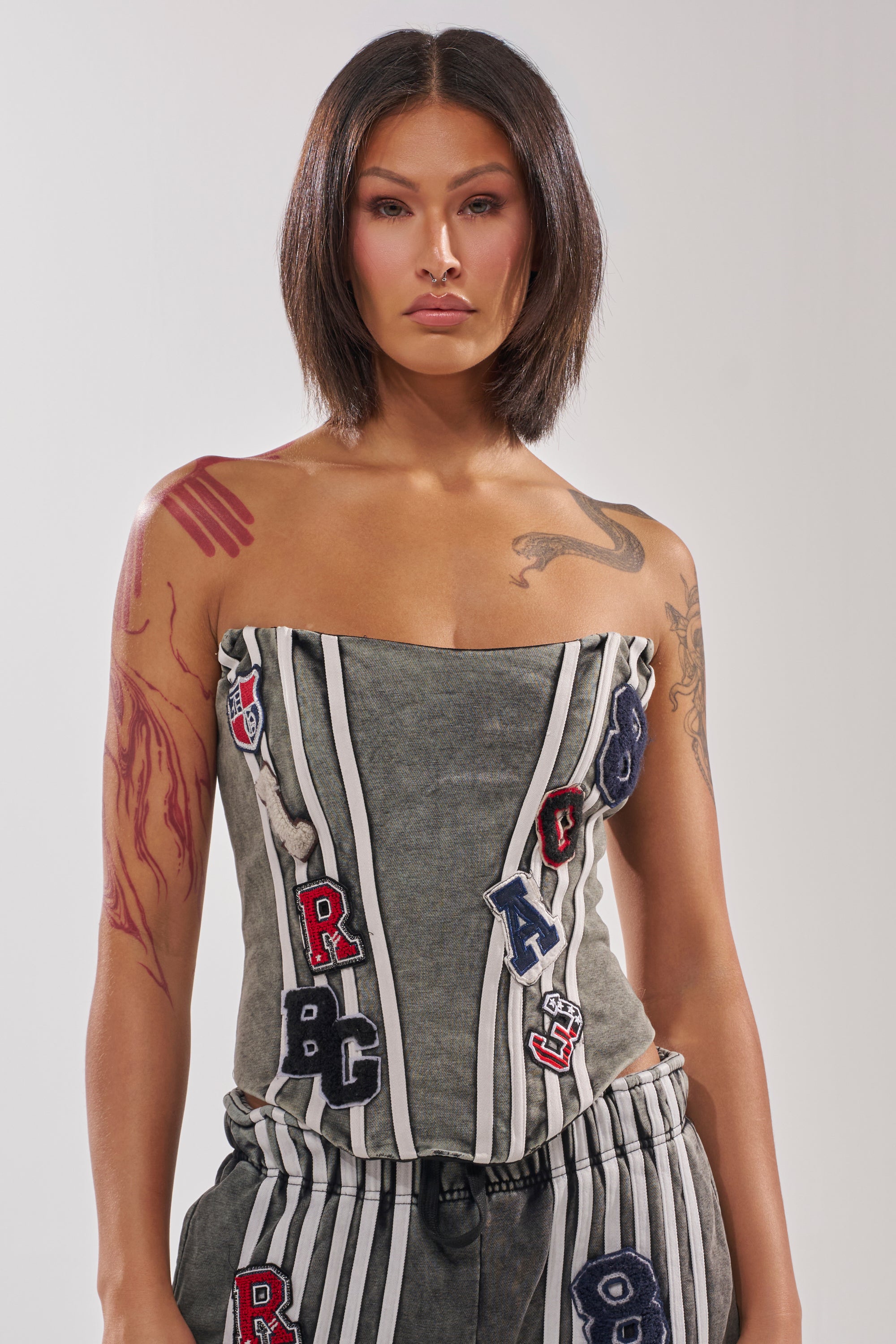 A woman with straight, shoulder-length dark hair wears the ROCK AND ROLL CORSET in gray with matching striped pants and visible arm tattoos, standing against a plain background. 1