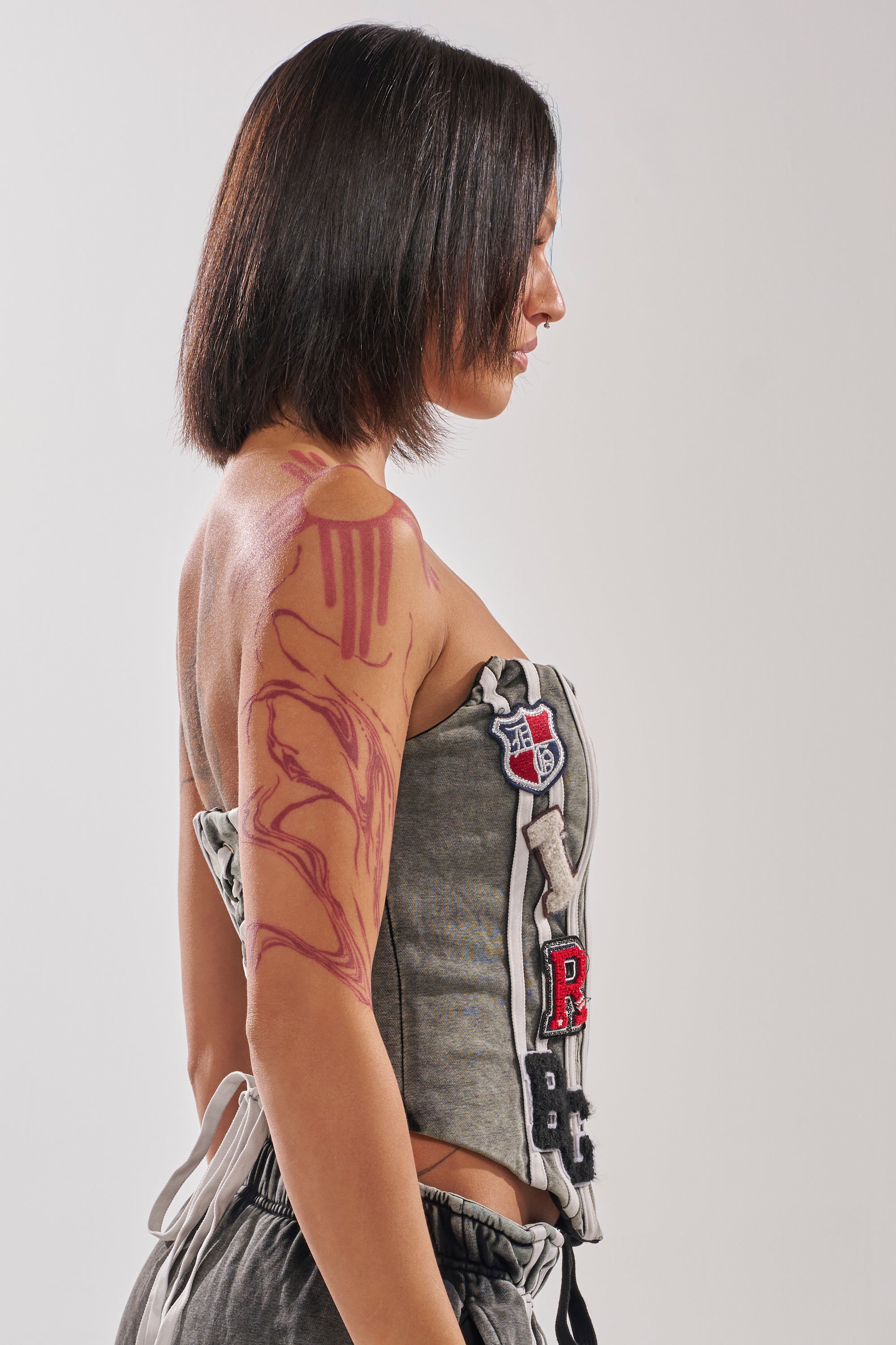 A woman with straight, shoulder-length hair stands in profile against a plain background, wearing the gray ROCK AND ROLL CORSET with lace-up back and patches, paired with sweatpants. A large pink abstract tattoo covers her left arm and shoulder. 3