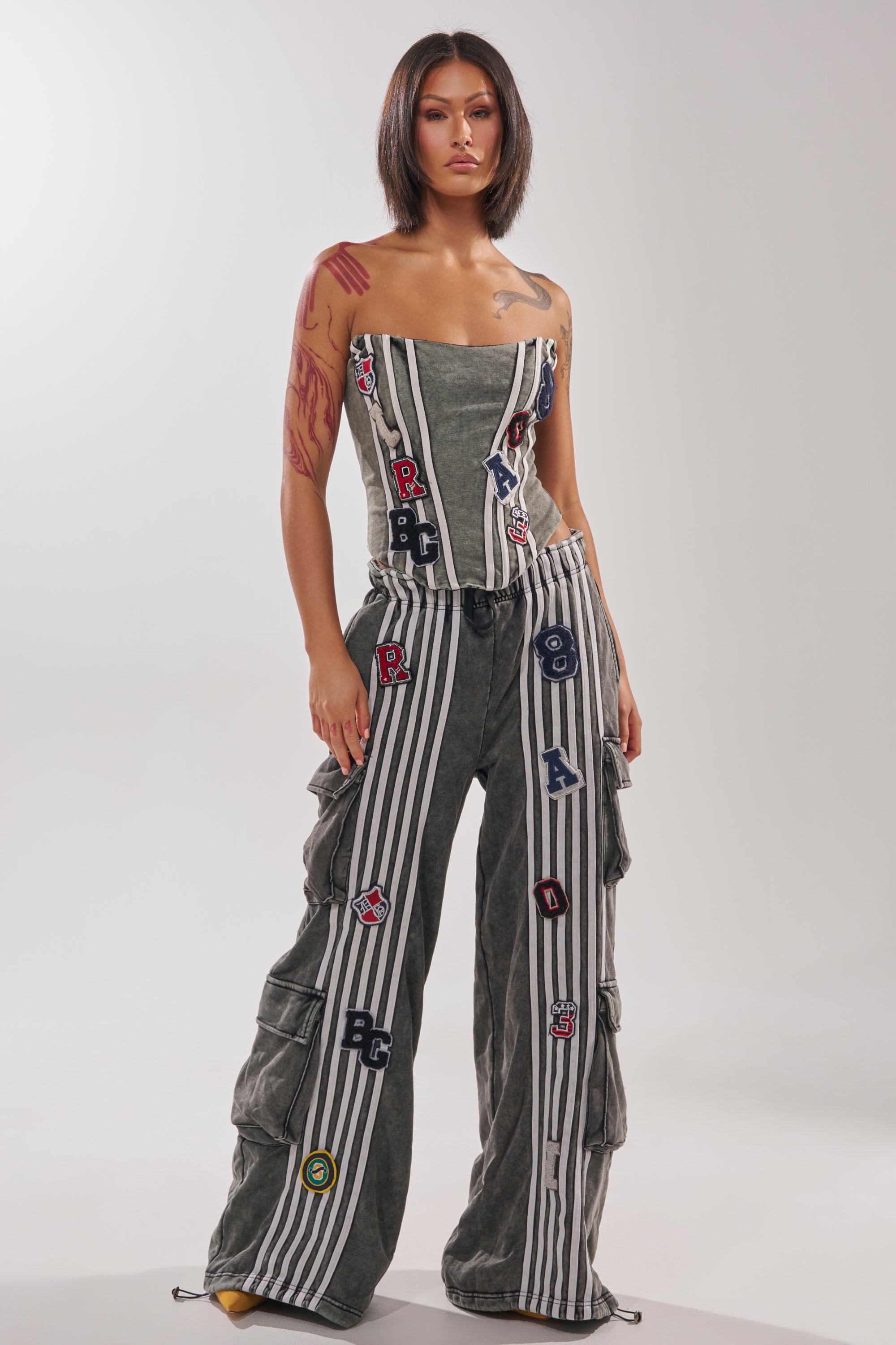 A woman with a bob haircut models the ROCK AND ROLL CORSET, a strapless striped jumpsuit with colorful letter patches and ruffle details, against a plain white background. 5