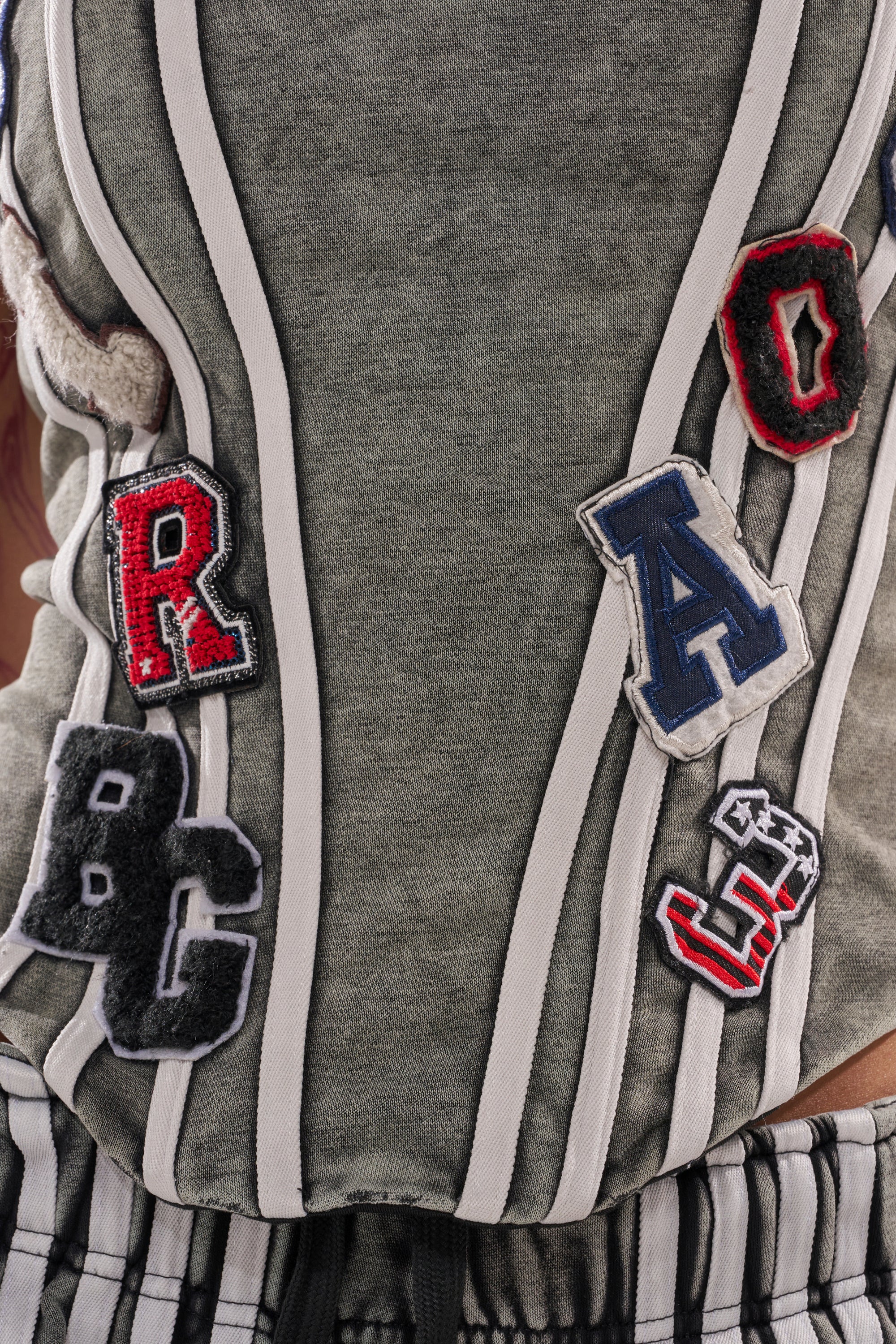 A close-up of the ROCK AND ROLL CORSET—a gray and white striped shirt with colorful varsity-style letter patches in red, blue, white, and black, finished with a bold lace-up back corset detail. 7