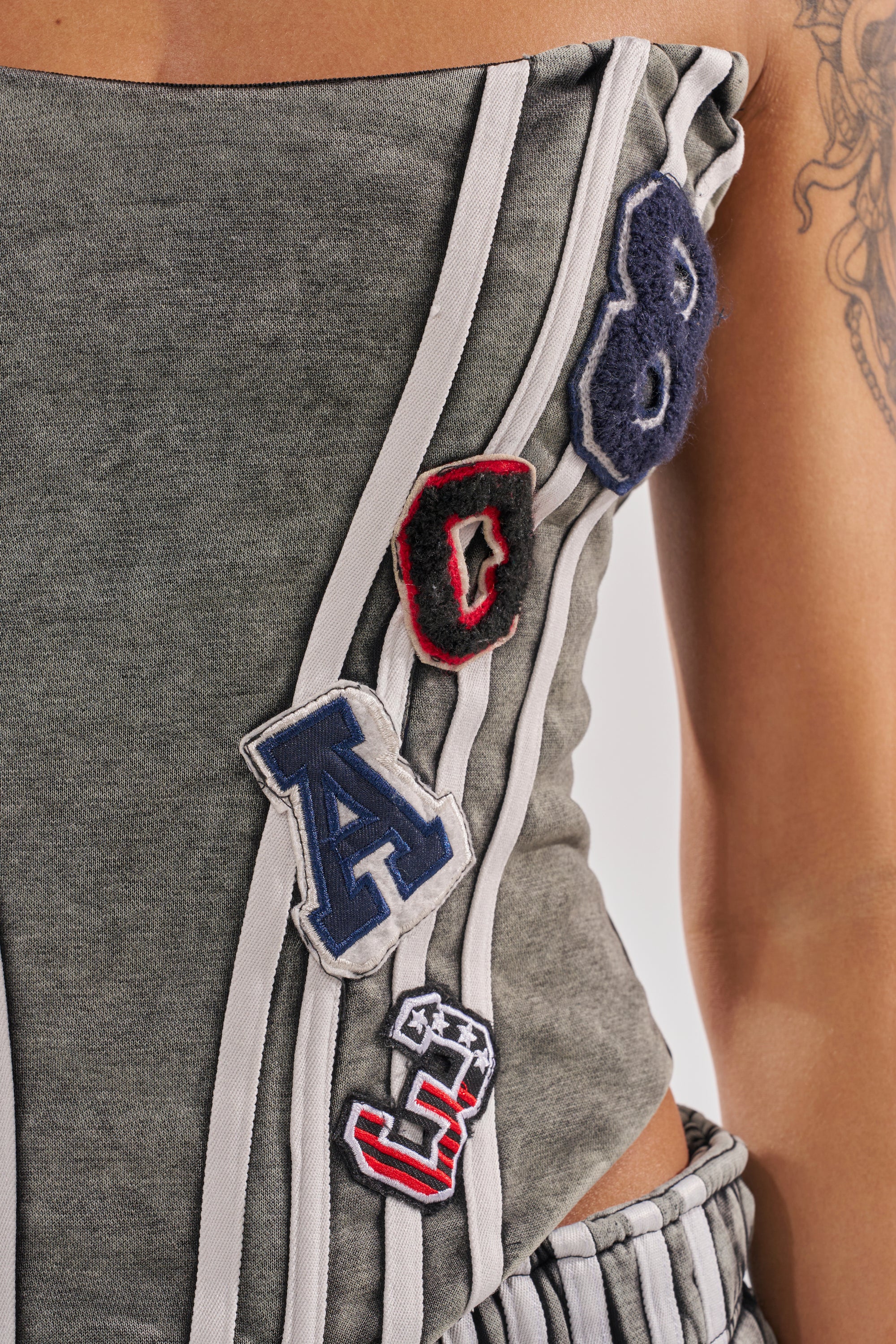 A close-up shows someone in the ROCK AND ROLL CORSET—a grey, striped, strapless top with letter and number patches (A, 8, 0, 5) sewn on. A visible arm tattoo adds an edgy touch to the look. 8