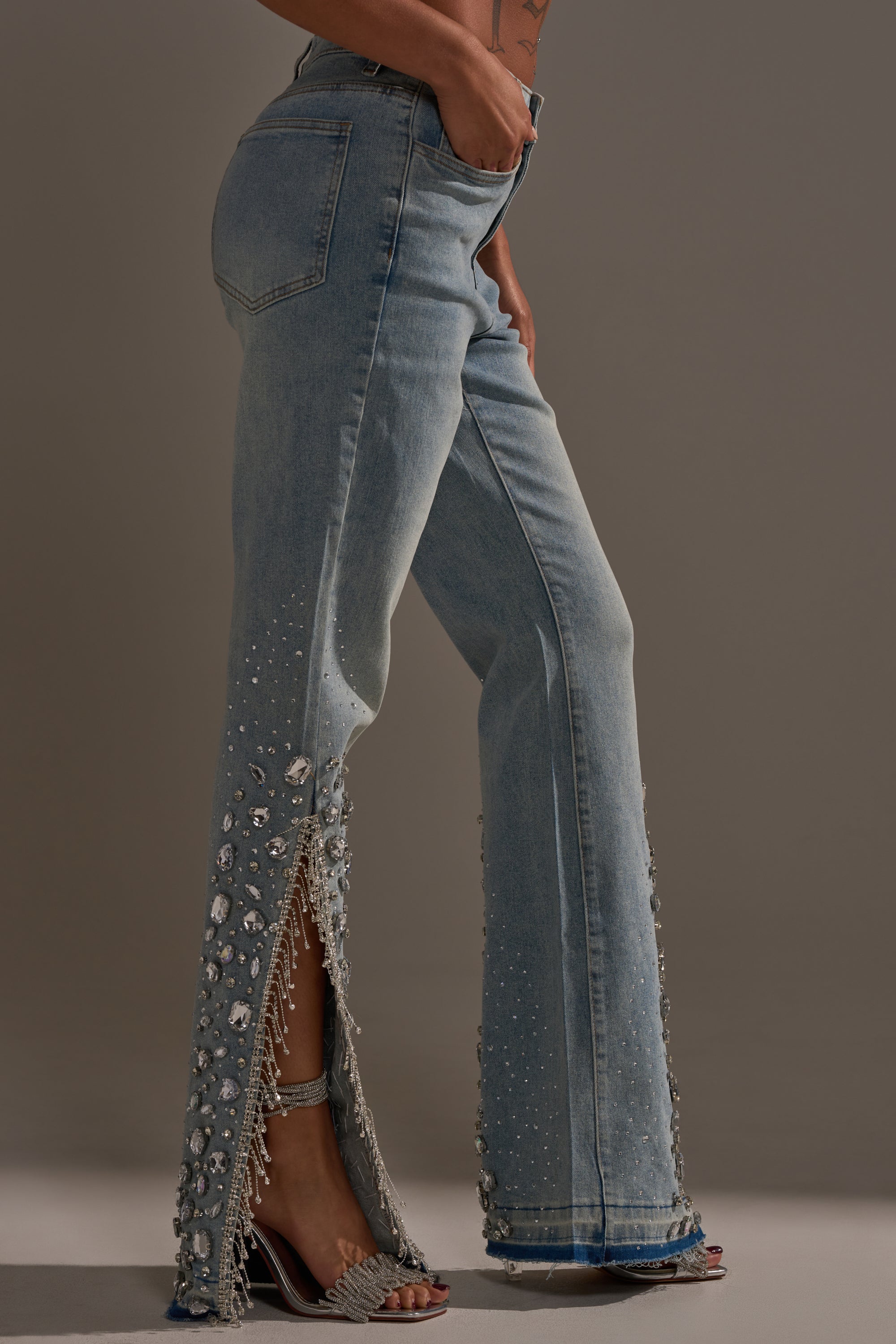A person models the STAR GIRL RHINESTONE DENIM—light blue, high-rise jeans adorned with pearls, sequins, and rhinestones, featuring side slits—paired with silver high-heeled sandals against a plain background.