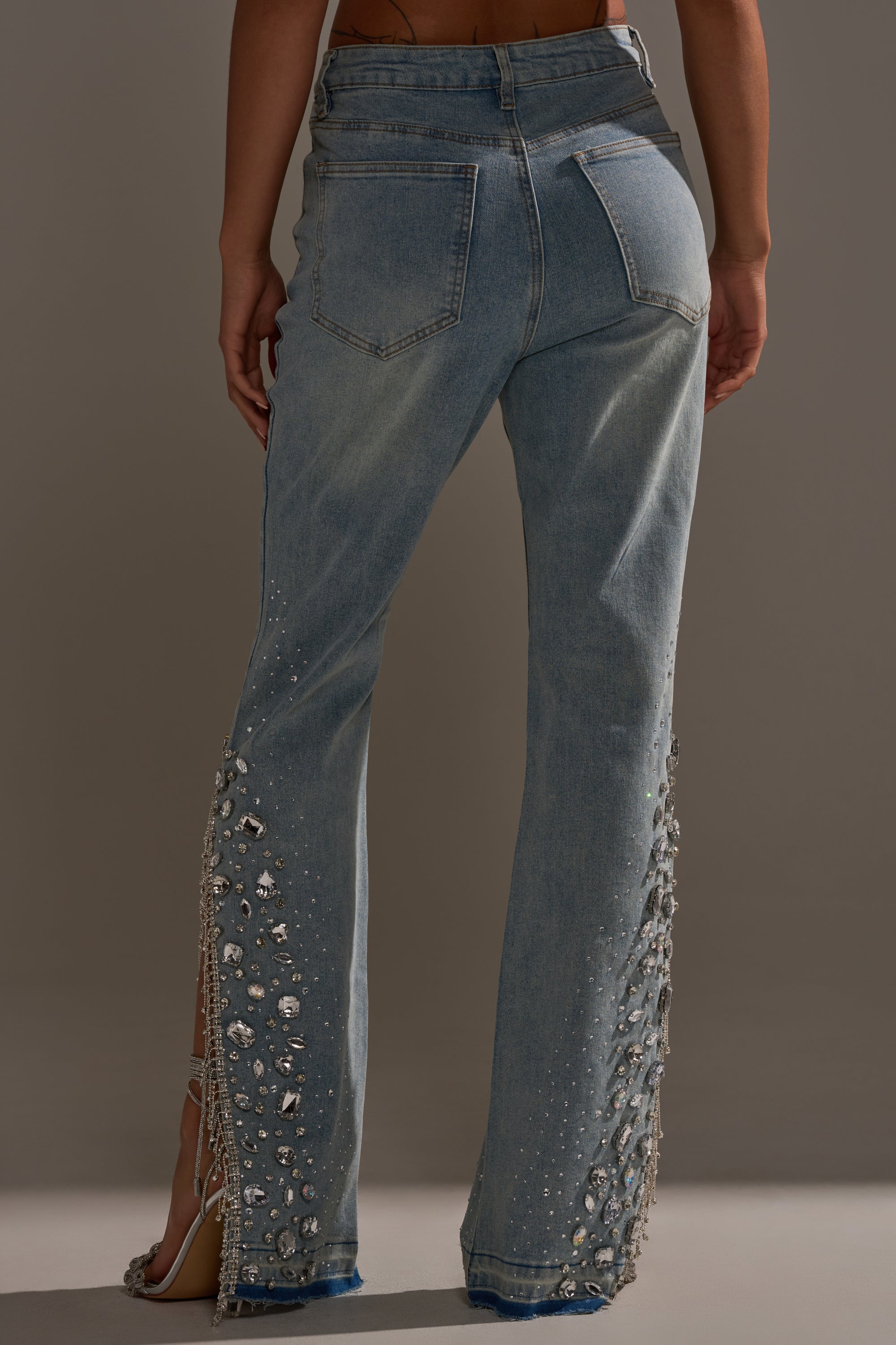A person shown from the back wearing STAR GIRL RHINESTONE DENIM—high-rise flared-leg jeans adorned with sparkling rhinestones and beads along the lower legs, featuring back pockets and a relaxed fit.