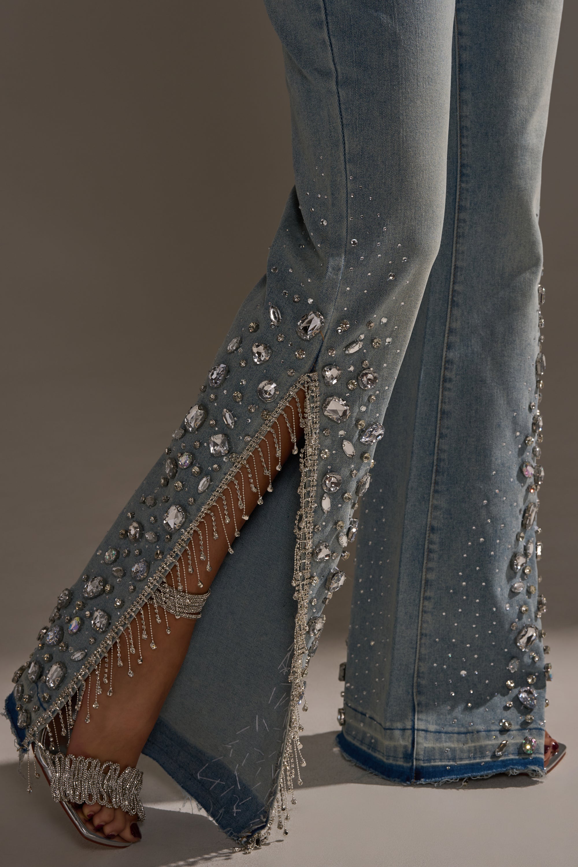 A close-up of a person in STAR GIRL RHINESTONE DENIM jeans with bead and fringe details on flared, split hems, revealing sparkly high-heeled sandals against a simple background.