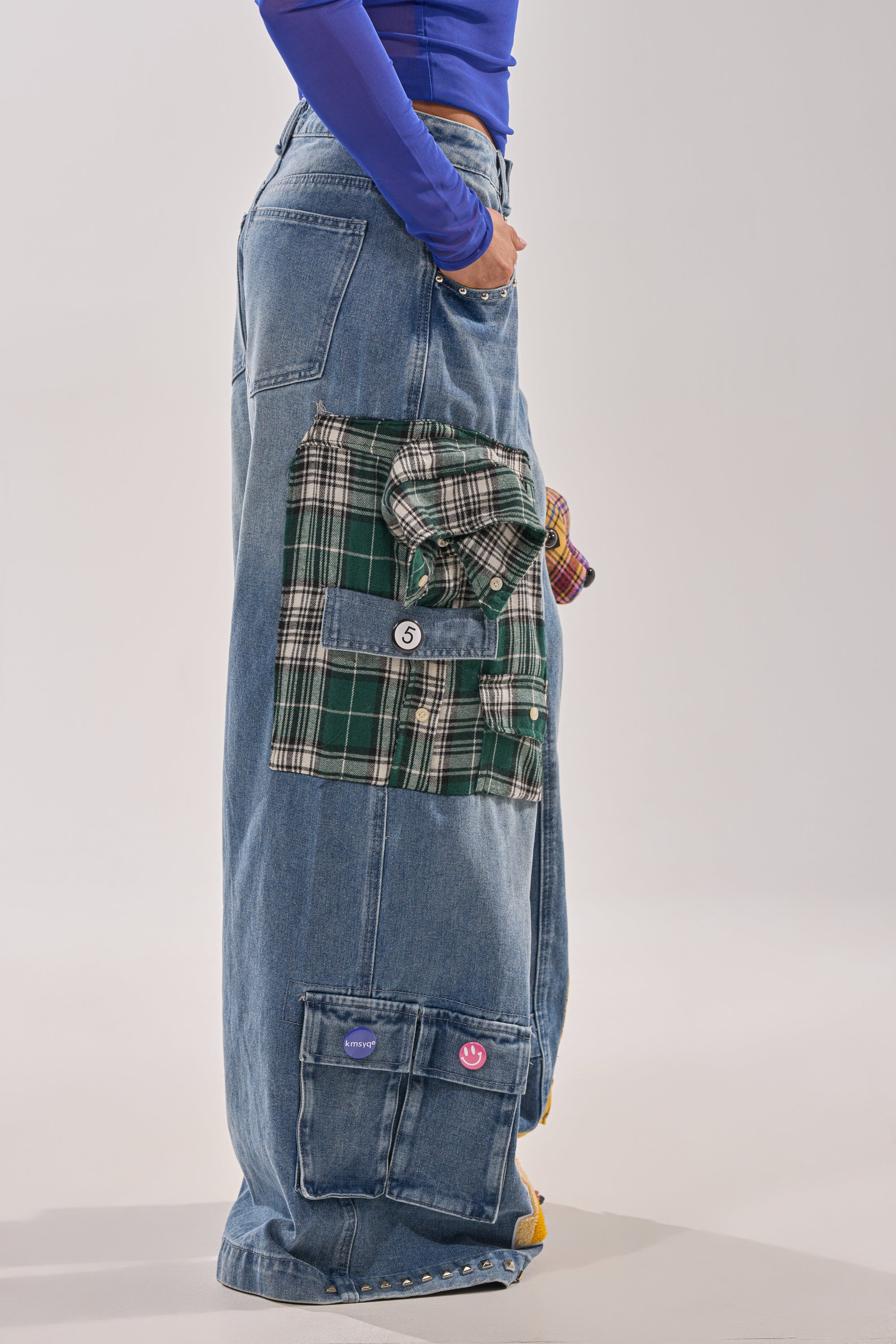 Someone stands in profile against a plain background, wearing FEELING THE WAVES RELAXED DENIM—wide-leg jeans with oversized cargo-style pockets, including a green plaid shirt pocket and a blue denim pocket. 2