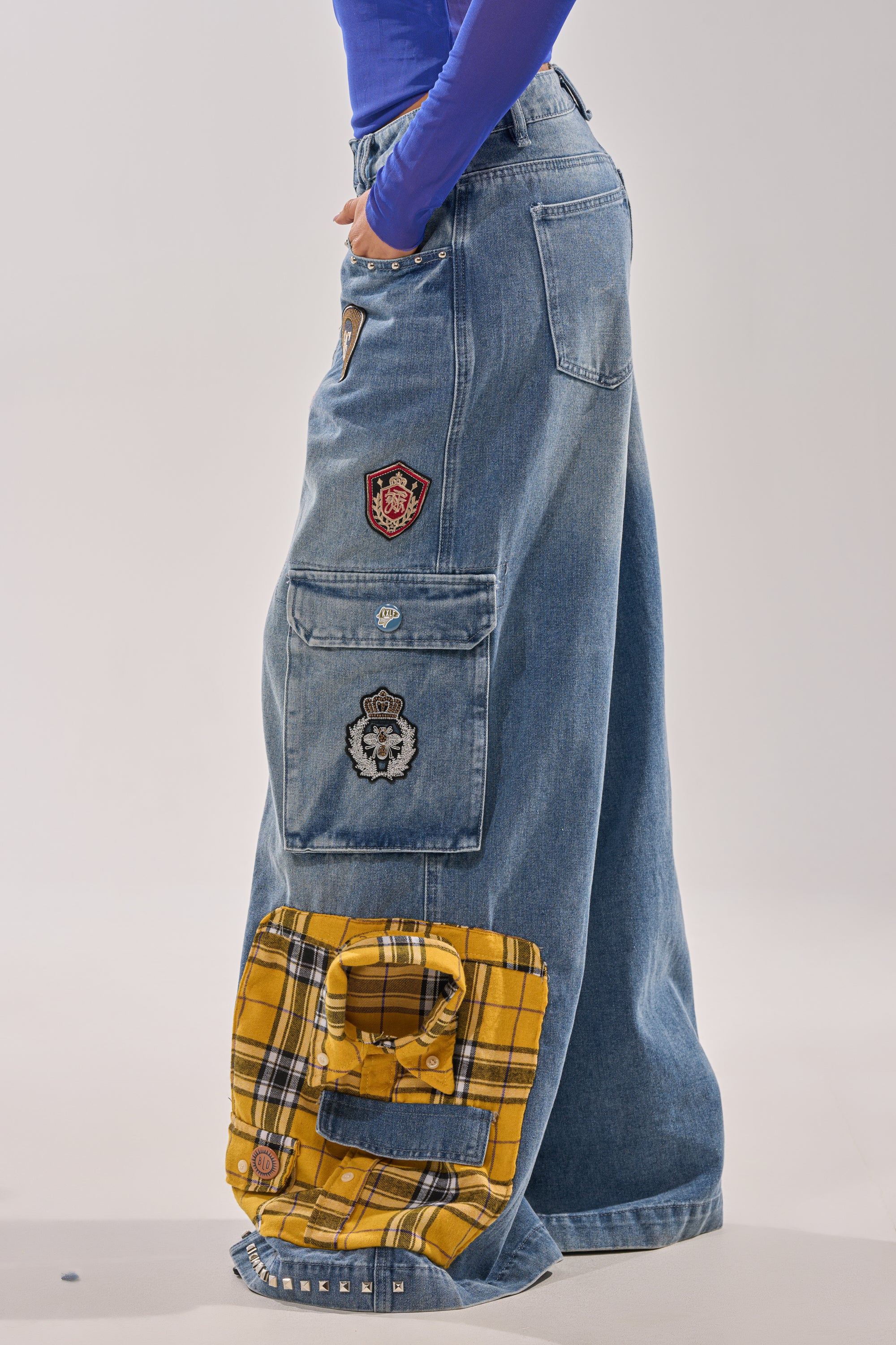 A person wearing the FEELING THE WAVES RELAXED DENIM, featuring wide legs, cargo pockets, decorative patches, and a yellow plaid detail near the hem, paired with a fitted blue long-sleeve top. 3