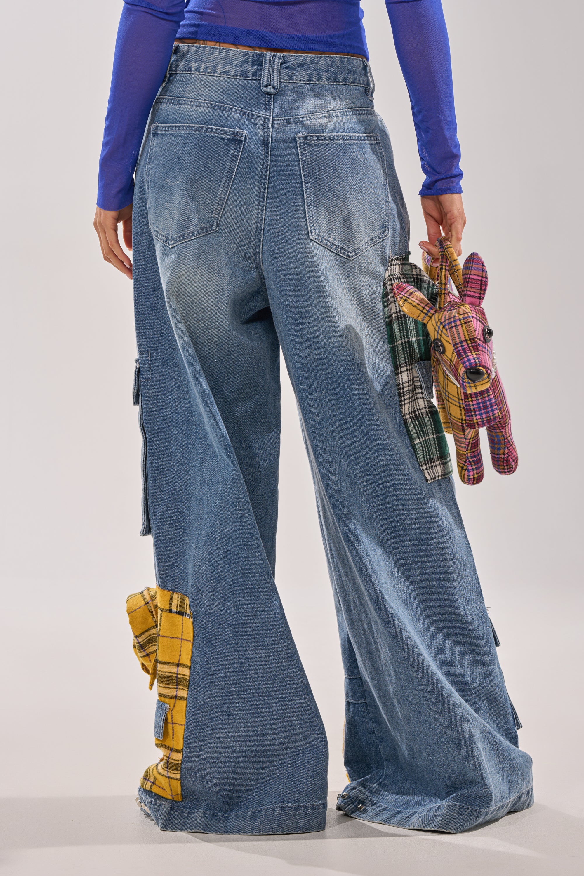 Wearing FEELING THE WAVES RELAXED DENIM with yellow plaid accents and a blue long-sleeve top, this person holds a small plaid stuffed dog and green plaid pouch. The behind-shot photo radiates an effortless streetwear vibe. 4