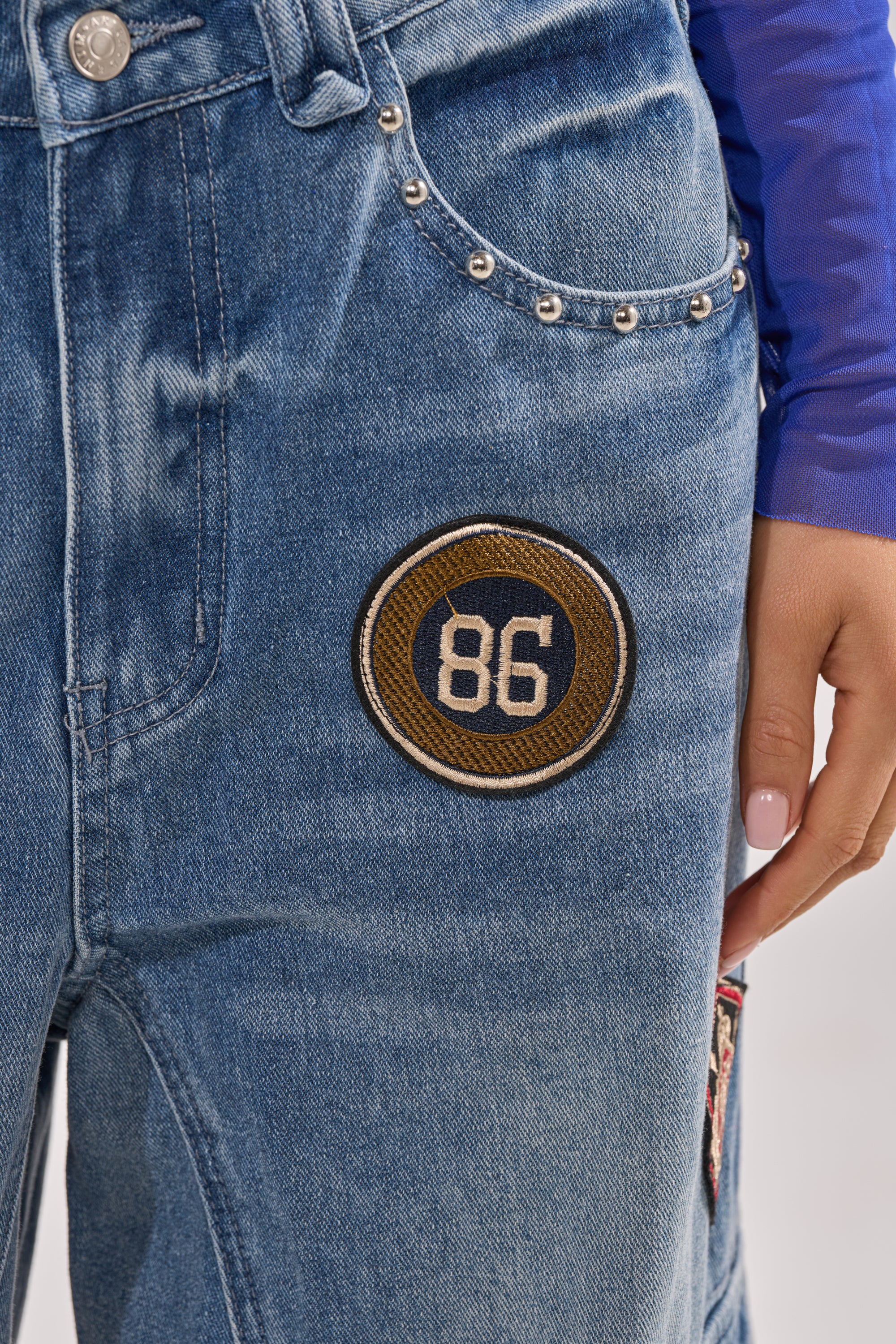 Close-up of the FEELING THE WAVES RELAXED DENIM featuring metal studded pockets, a yellow-bordered "86" patch, cargo-style pockets, and a hand with pale pink nail polish resting on the blue wide-leg jeans. 6