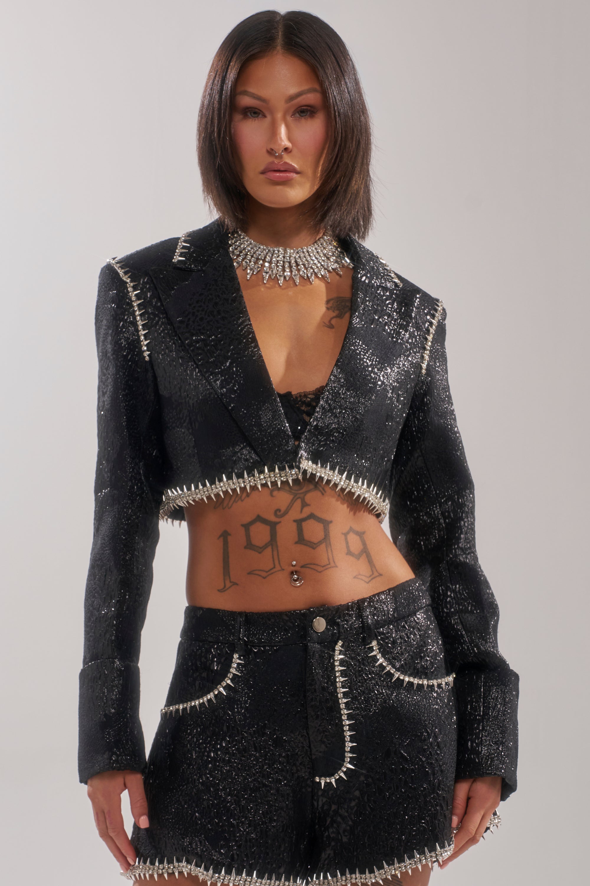 A person with straight dark hair wears the TWILIGHT ZONE EMBELLISHED BROCADE BLAZER, featuring sparkly black fabric and rhinestone spike trim, paired with matching shorts, a spiked necklace, and a visible "1999" stomach tattoo. 0