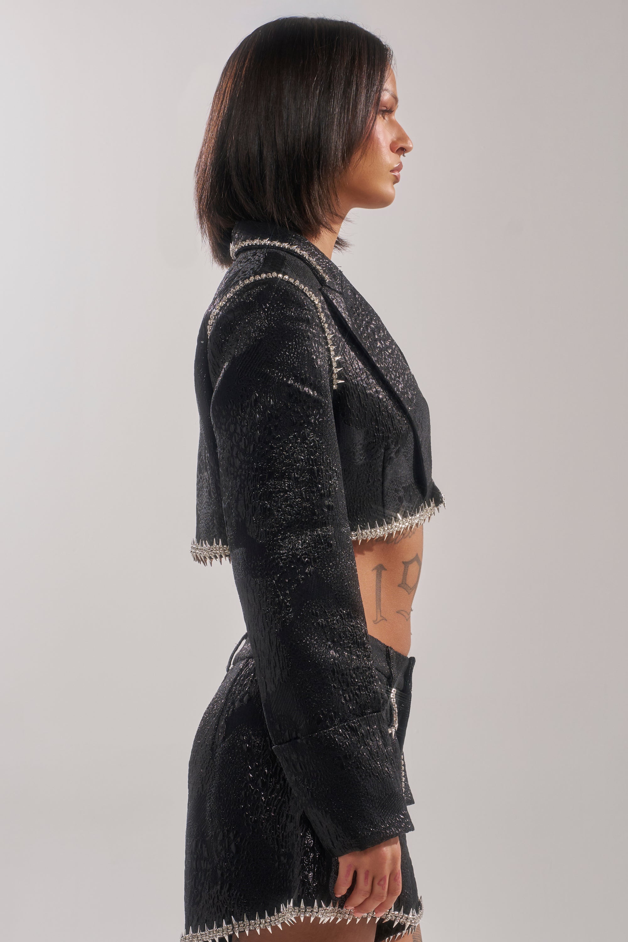 A woman with straight dark hair stands in profile, wearing the TWILIGHT ZONE EMBELLISHED BROCADE BLAZER and matching high-waisted shorts, revealing a side tattoo. The background is plain and light-colored. 5