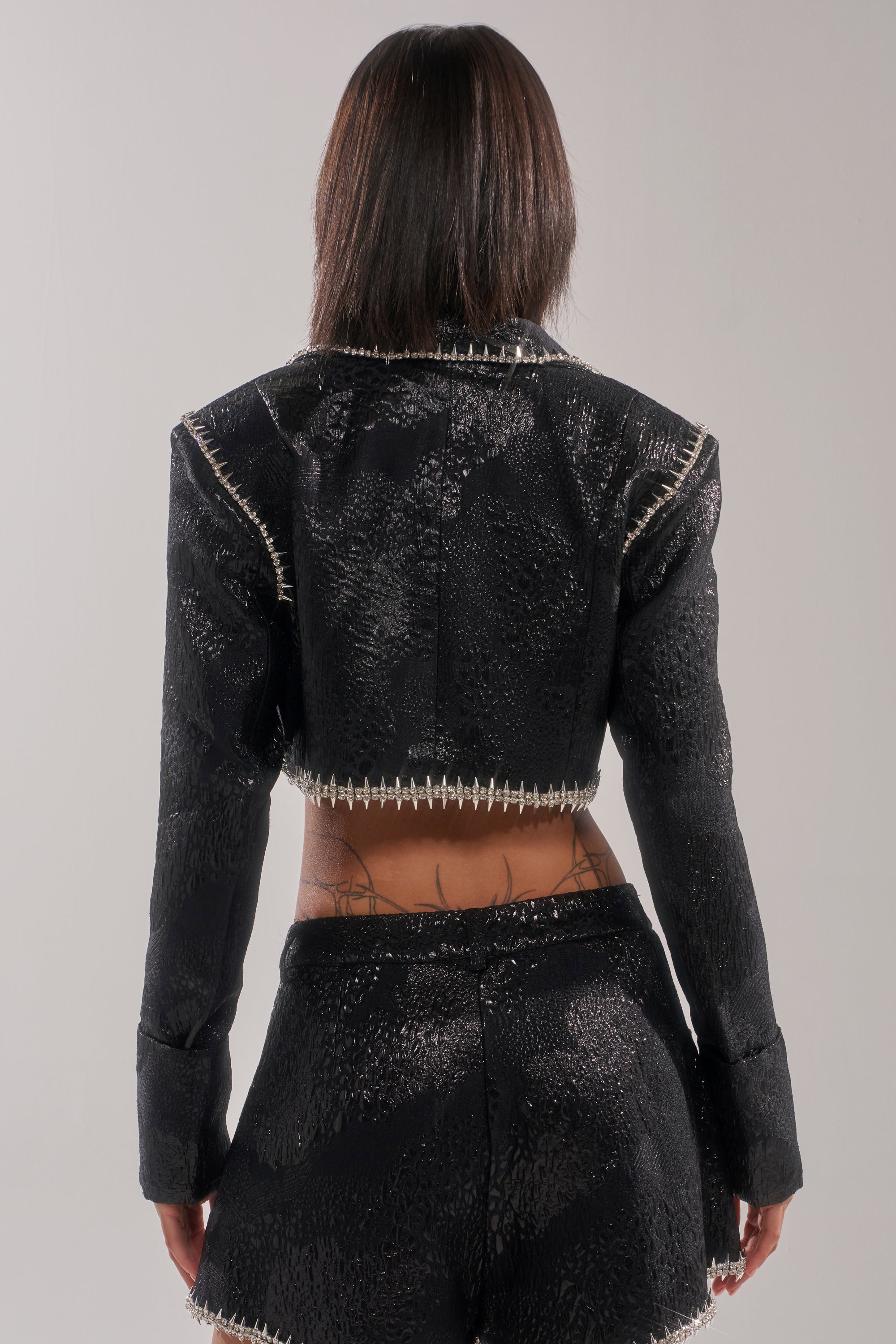 A person with straight shoulder-length hair is shown from the back, wearing the TWILIGHT ZONE EMBELLISHED BROCADE BLAZER and matching high-waisted shorts with rhinestone spike trim, revealing lower back tattoos. 6