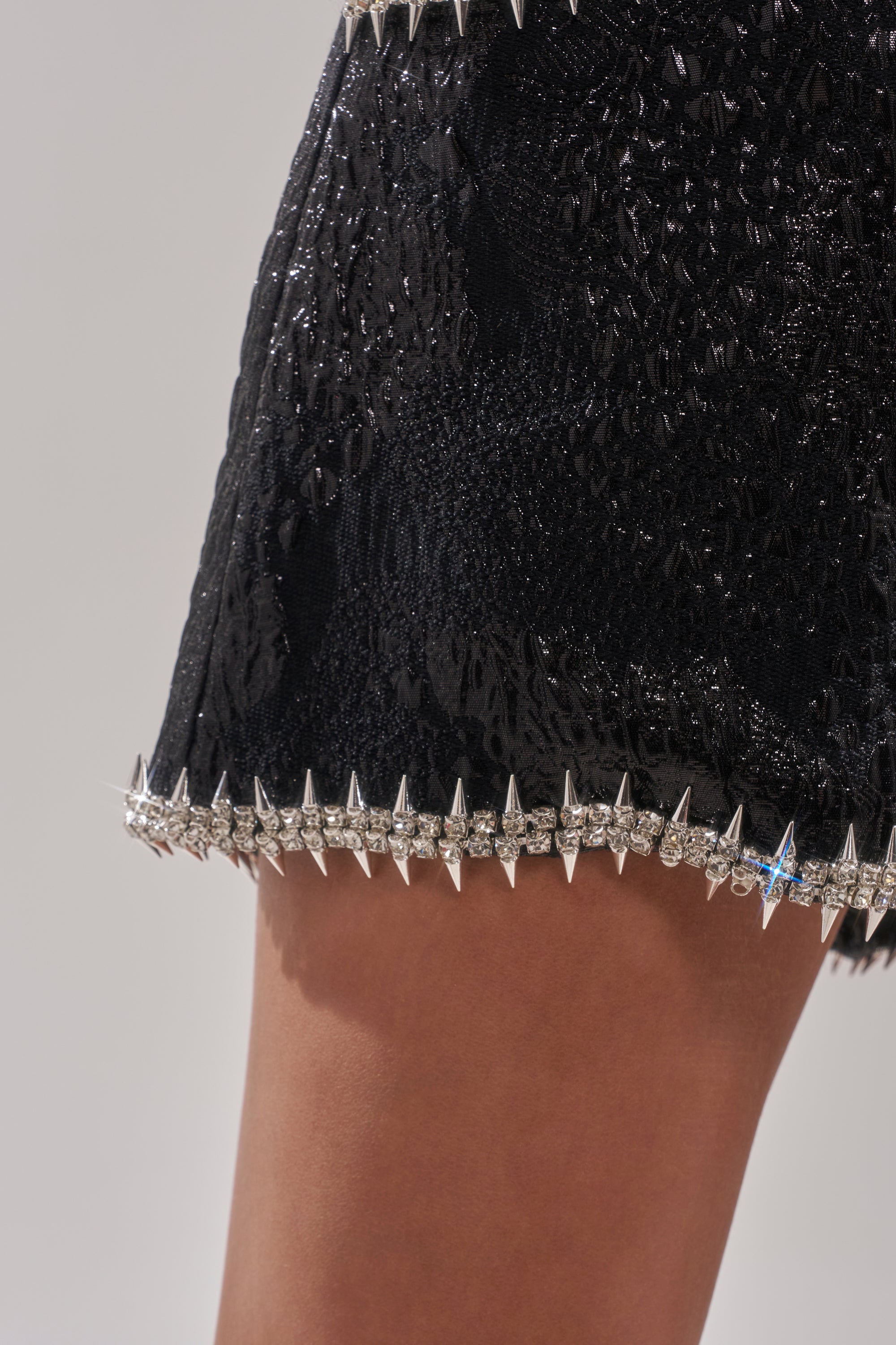 A close-up of a person wearing the TWILIGHT ZONE EMBELLISHED BROCADE SHORTS, featuring black textured fabric with a border of silver spikes and clear rhinestones along the hem for an edgy rocker vibe. 8